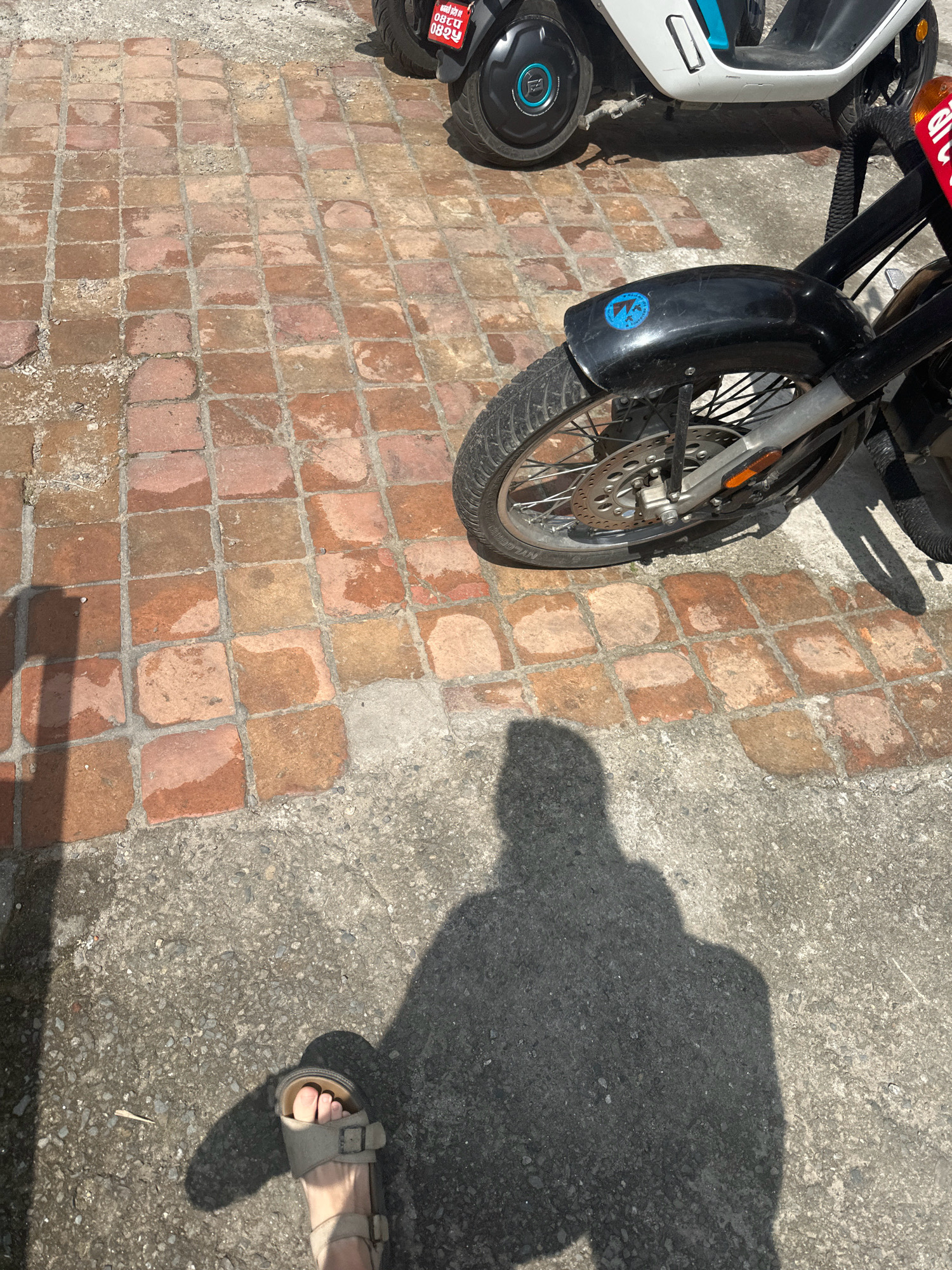 In Lalitpur, Nepal, on a paved area of red brick squares and rough concrete, a person's sandaled right foot is visible in the foreground, casting a shadow. Two motorcycles are present; one in the foreground is black with a blue sticker on the fender and a red and white license plate. Partially visible behind it is a white and blue electric scooter. The lighting suggests it is daytime.
