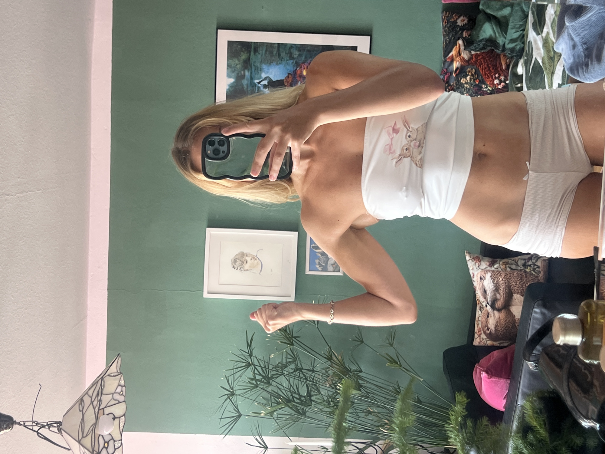 An individual is taking a mirror selfie in a room. The person is wearing a white strapless top with a rabbit graphic and white shorts. They are holding a phone to obscure their face. In the background, there is a green wall with framed pictures, a plant, and a couch with pillows. The room appears to be a bedroom or living space, possibly in a residence. There is no indication of the specific location within Mühldorf a. Inn, Germany.