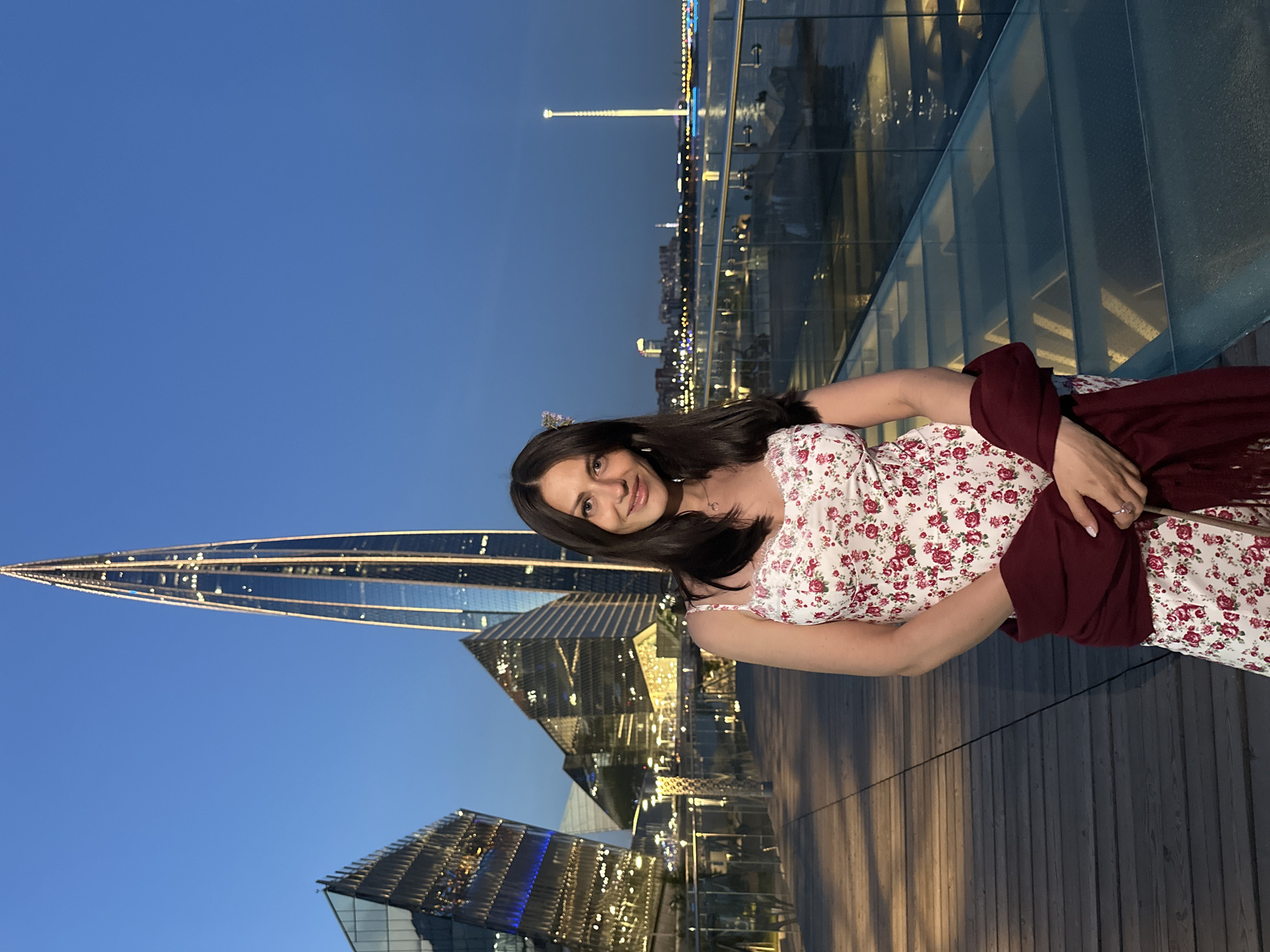 The image shows a woman standing on a wooden walkway at dusk with modern glass buildings and a tall, illuminated skyscraper in the background. The woman is wearing a white floral dress and a dark red shawl draped over her shoulders. She is smiling and looking towards the camera. The architectural style of the buildings, particularly the prominent skyscraper, suggests the location is the Lakhta Center in Saint Petersburg, Russia. The scene is set against a clear blue twilight sky.