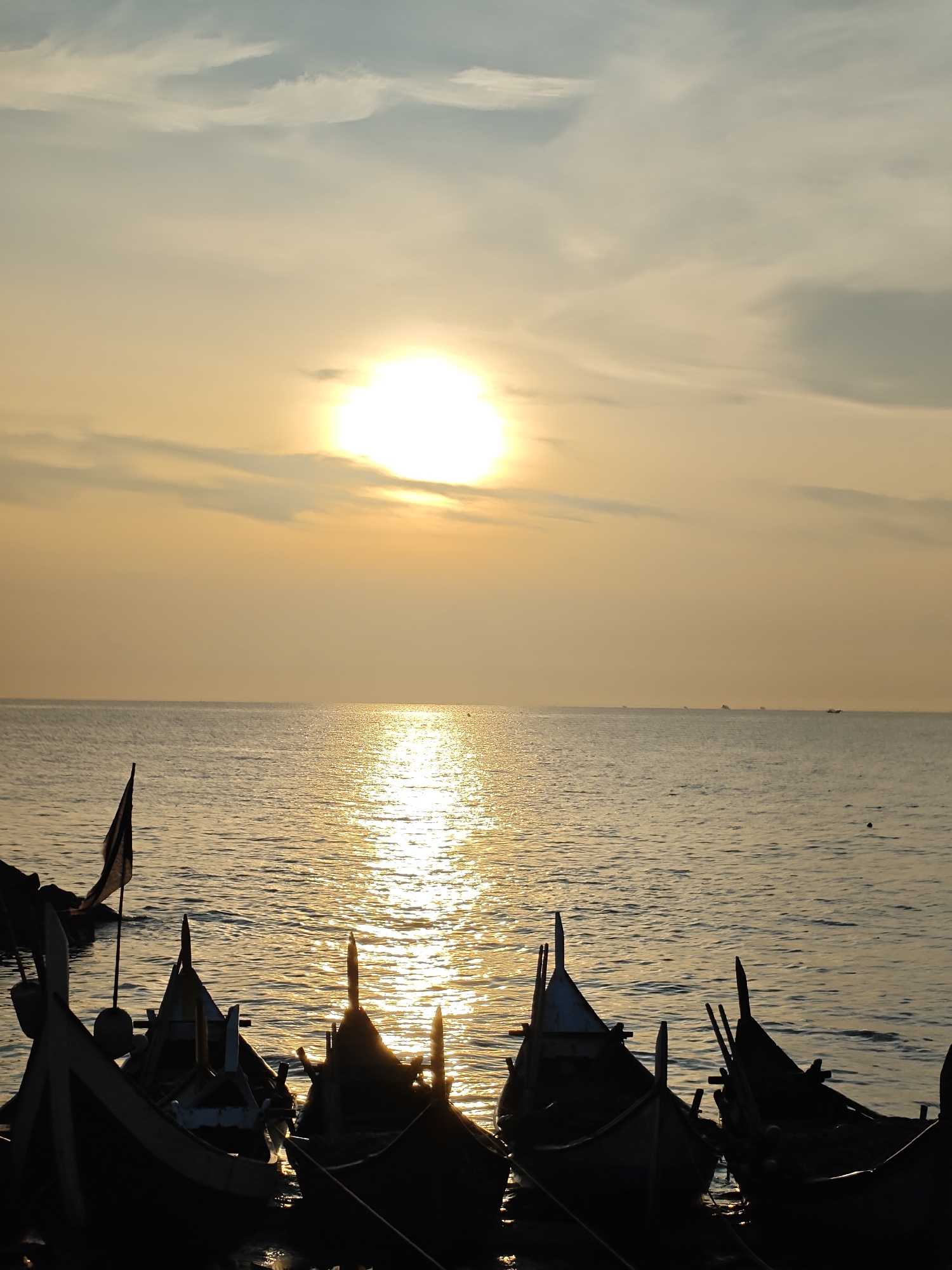 The image depicts a coastal scene at sunset or sunrise in Lhokseumawe, Indonesia. The sky is bright, with the sun appearing as a diffused, bright spot obscured by thin clouds. The ocean reflects the sunlight, creating a shimmering path toward the horizon. Several small fishing boats are silhouetted in the foreground, docked or moored near the shore. A flag is visible on one of the boats. In the distance, there are faint outlines of what appear to be additional vessels on the horizon.