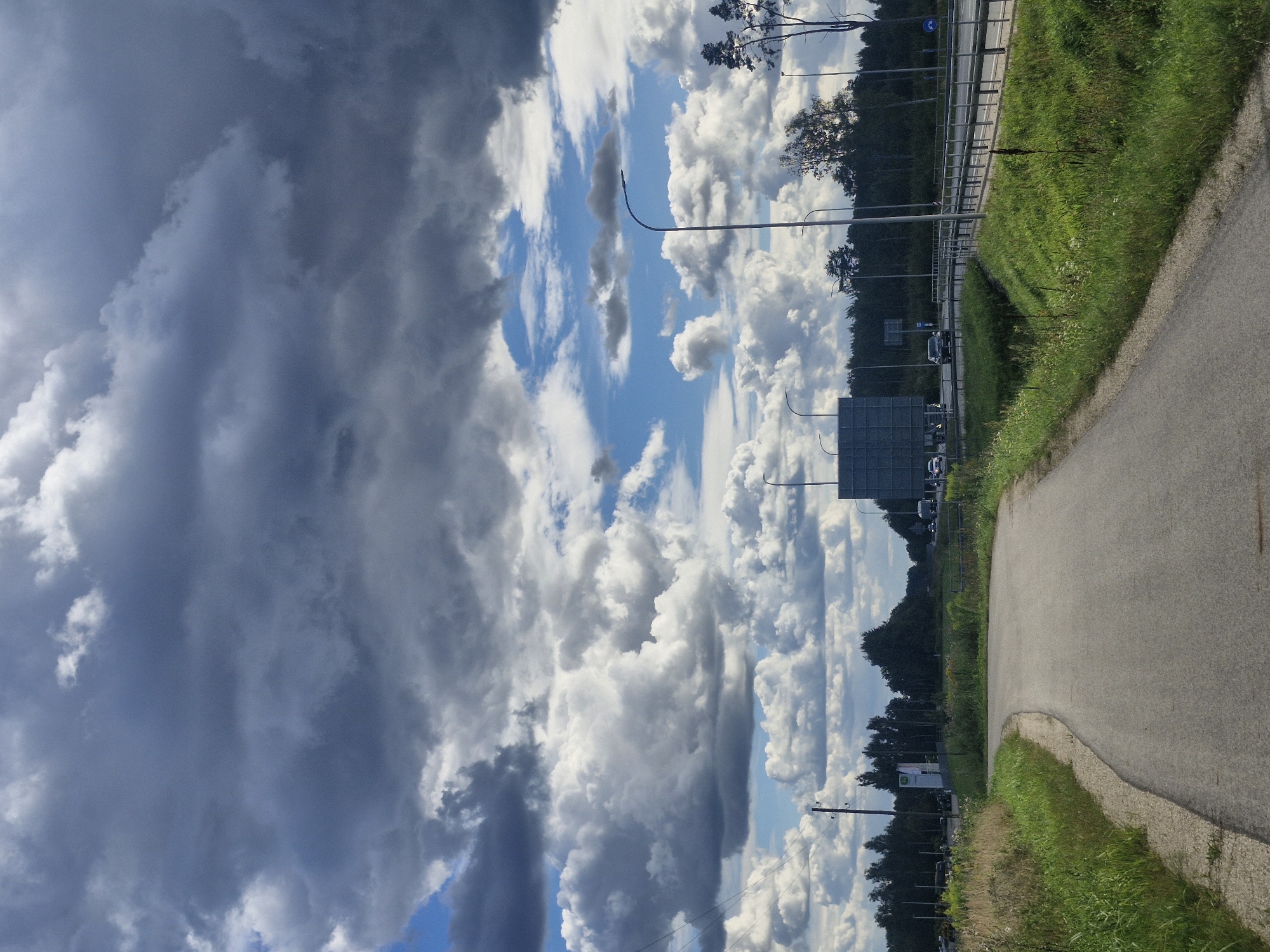 The image depicts a road curving to the left, with a grassy verge on either side. In the background, a dense line of trees marks the horizon. Beyond the trees, a highway with a metal guardrail is visible, with several cars driving on it. Streetlights are positioned along the highway. The sky is filled with dramatic, dark clouds, interspersed with patches of bright blue sky and large, fluffy white cumulus clouds. The scene appears to be a rural or semi-rural area in Latvia.