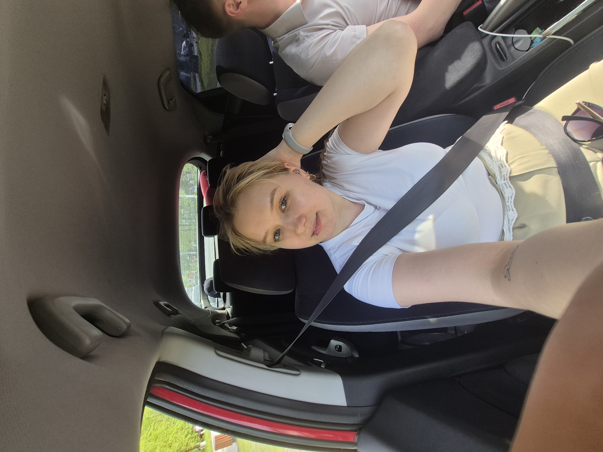 A woman wearing a white t-shirt and seatbelt is in the passenger seat of a car. Another person, wearing a light-colored shirt, is in the backseat. The car's interior is visible, including the roof grab handle. Outside the car, greenery and a building are visible through the window. The location may be Chaikovsky, Russia, though no specific landmarks are definitively visible.