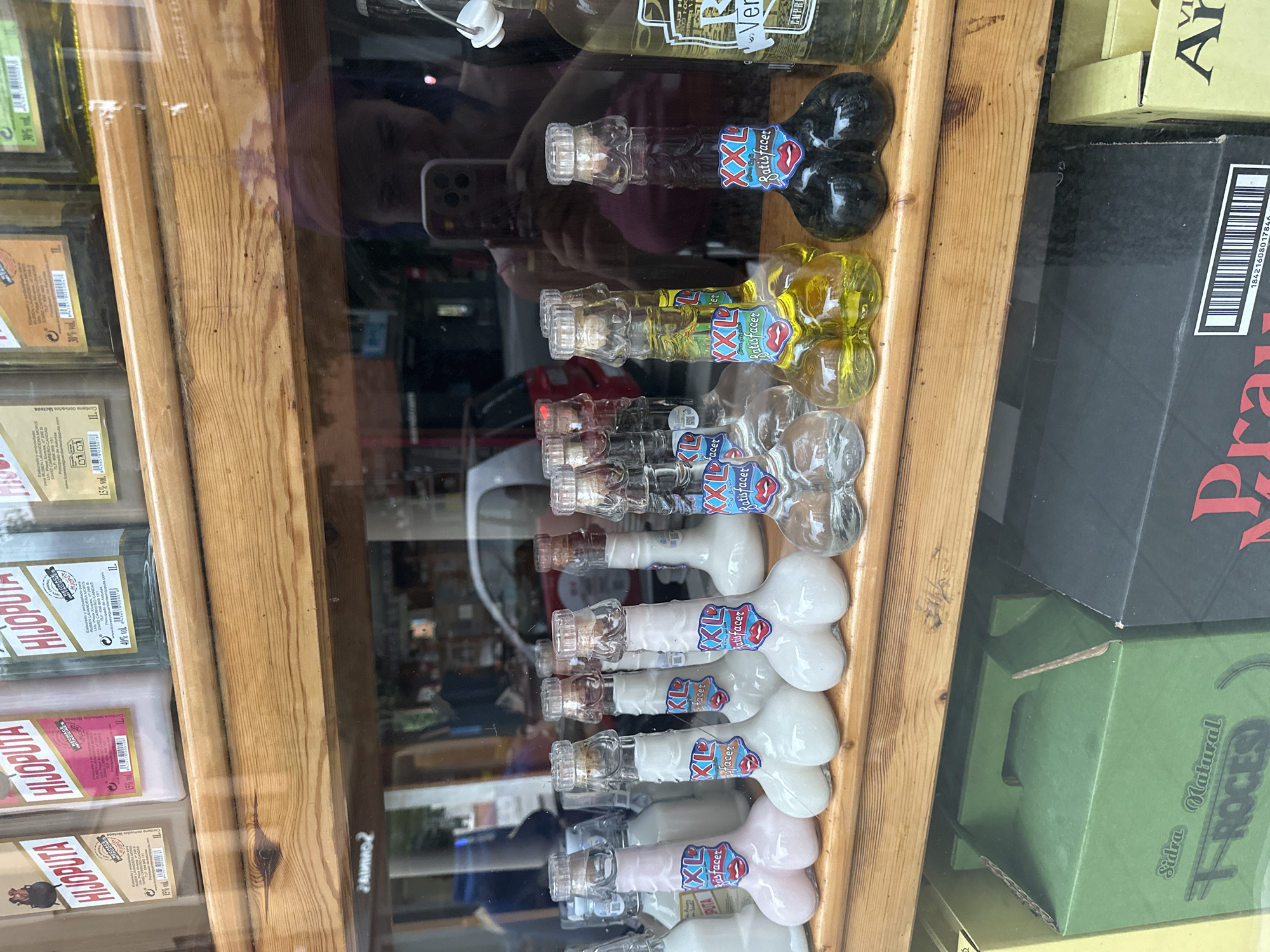 The image shows a display in a shop window. The shop is located in Gijón/Xixón, Spain, based on the "Playa Españoles" signage. The display features small, novelty bottles shaped like penises, filled with various colored liquids (black, yellow, clear, red, pink). The bottles are labeled "XXL Satisfacer" or "XL Satisfacer." Other products, including bottles labeled "HIJOPUTA," are also visible on the left side of the frame. To the right are boxes, with partial text reading "Sidra Natural ROCES" and "Pra." A person's reflection, including their phone, is visible in the shop window. The display is surrounded by a wooden frame.