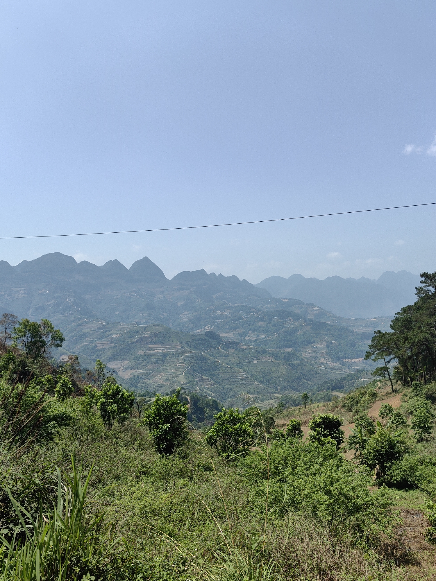 The image presents a panoramic view of the mountainous landscape in Bạch Đích Commune, Vietnam. In the foreground, dense green vegetation covers the lower hills. The middle ground reveals terraced fields carved into the slopes, with scattered settlements visible. Multiple mountain ranges are visible in the distance, fading into a light blue sky. An overhead wire bisects the upper part of the frame.