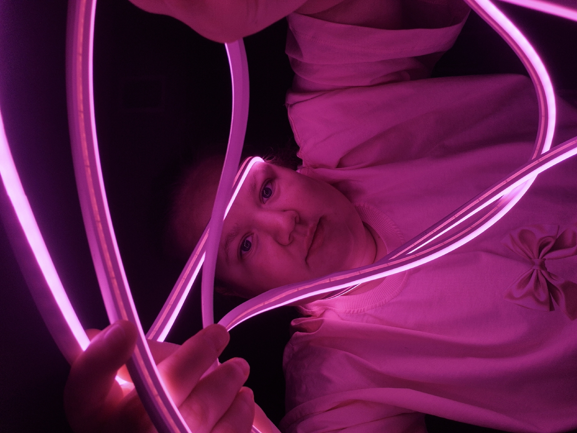 A person with fair skin and blue eyes is holding pink neon lights in front of their face. They are wearing a pink shirt with a bow on the right shoulder. The background is dark and out of focus. The location is unknown and not inferable from the image.