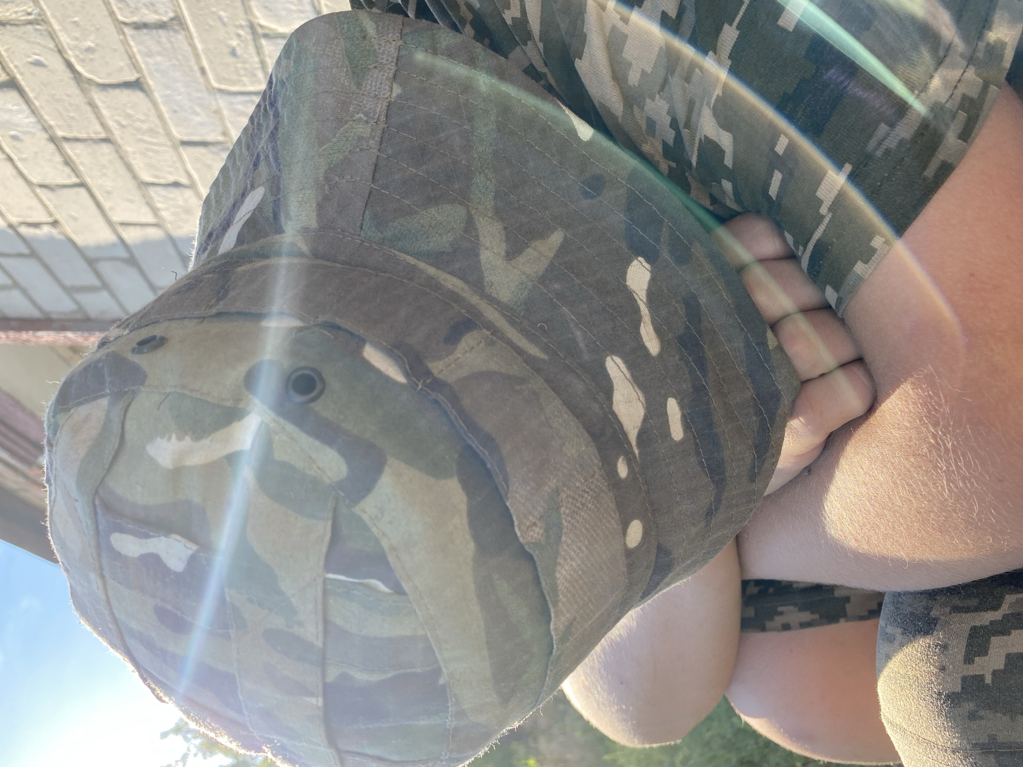 The image shows a close-up of a person wearing camouflage clothing, including a bucket hat and a shirt. The subject's arm and hand are visible, as well as part of their leg. The camouflage pattern is a mix of greens, browns, and tans. The lighting suggests it is daytime, possibly sunny, with a lens flare visible. In the background, there is a hint of a tiled surface and some greenery. The scene is outdoors, and the orientation of the image is rotated. There is no visible text. Based on the provided location context, the setting is Nova Praha, Ukraine. The image doesn't provide enough information to determine the specific activity or event taking place or any discernible emotions.