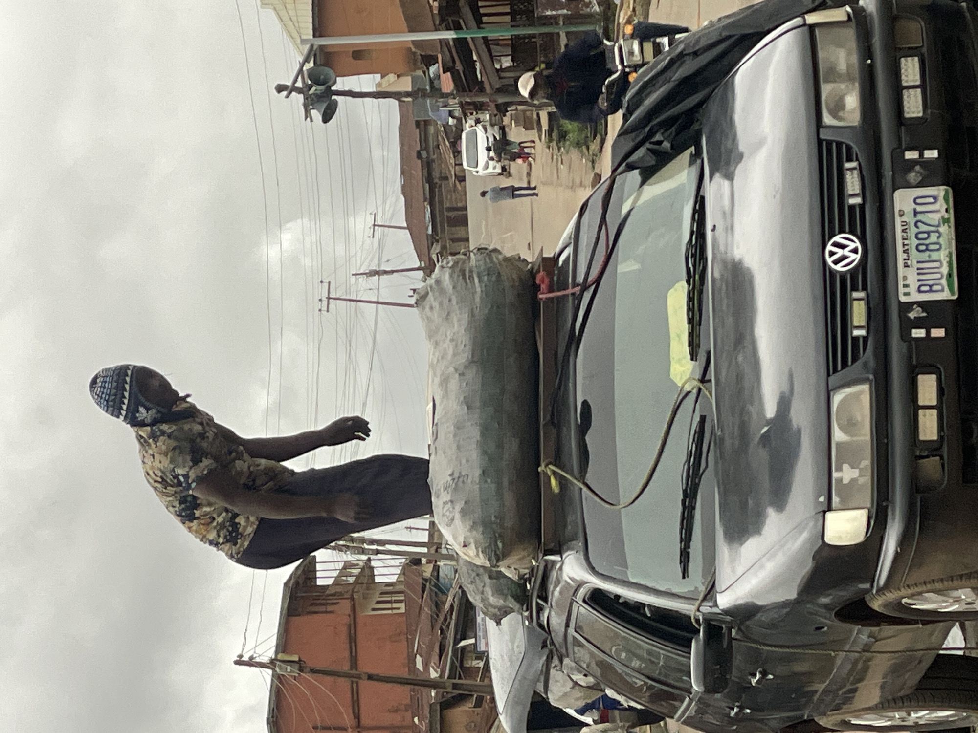 This media file is an outdoor street scene, rotated 90 degrees counter-clockwise, showing a man and a vehicle appearing vertically oriented under an overcast sky.
The primary subject is a dark-skinned man on the left, depicted as if standing upright against the sky, but vertically aligned with the image's rotated frame. He wears a patterned short-sleeved shirt, dark trousers, and a patterned head covering. His left arm is bent, and his right arm is outstretched.
To the right of the man is a dark-colored Volkswagen minivan or station wagon, also appearing vertically in the frame. The vehicle's front grille prominently displays the Volkswagen emblem. A Nigerian license plate is affixed to the front bumper, reading "PLATEAU BUU-892TO". Large, grey, cylindrical bundles are securely strapped to the roof of the vehicle, partially covered by a black tarpaulin or fabric. Ropes or straps are visible, securing the cargo.
In the background, behind the vehicle and man, is a street scene under a grey, cloudy sky, indicative of an overcast day. Utility poles with numerous crisscrossing wires are visible, along with parts of buildings that suggest a dense, urban or semi-urban environment. Further down the street, smaller figures of people and a white vehicle can be discerned. On the upper right, a structure with what appears to be loudspeakers or sirens is mounted on a pole or building.
The scene suggests activity related to travel or transport of goods, with the vehicle heavily loaded. The location is confirmed as Plateau State, Nigeria, by the license plate, aligning with the provided context of Jos, Nigeria. No specific emotions are discernible, and the overall mood is mundane and observational.