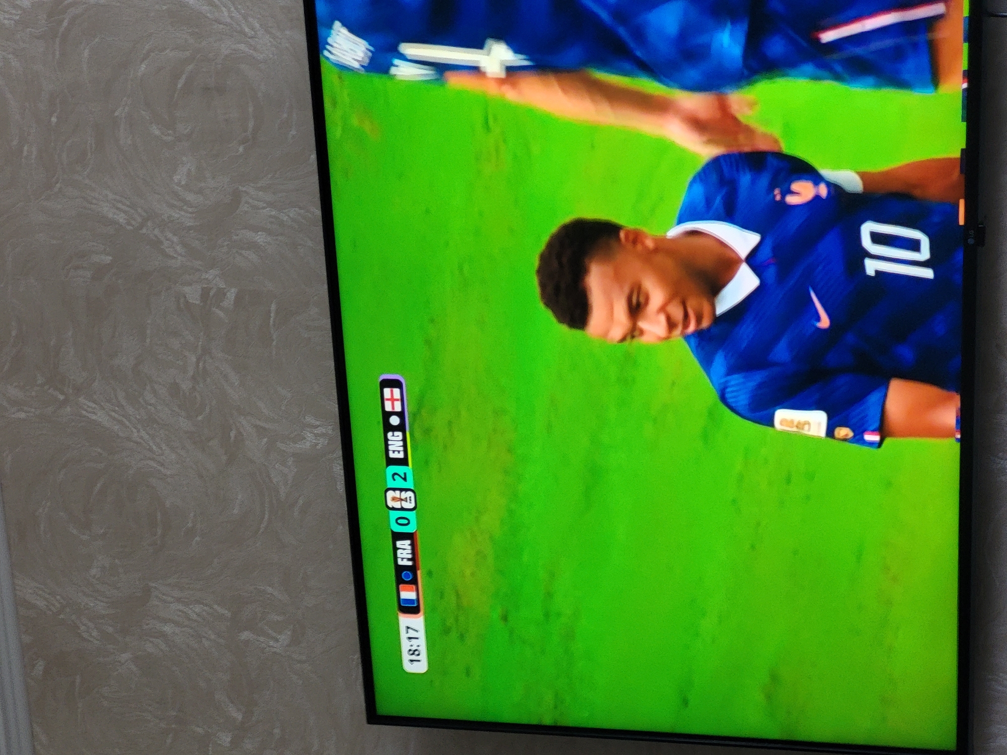 The image is a photograph of a television screen displaying a soccer match. The screen shows a score overlay indicating "FRA 0 - 2 ENG" at 18:17. Two soccer players are visible on the screen, both wearing blue jerseys. The player on the left, identified as Kylian Mbappé, is wearing the number 10. The player on the right is partially visible. The background on the screen is a vibrant green, representing the soccer field. The television is placed in front of a wall with a textured, light-colored wallpaper. The image does not contain any specific information or visual cues that would allow for identification of its location in the city of Petropavl, Kazakhstan.