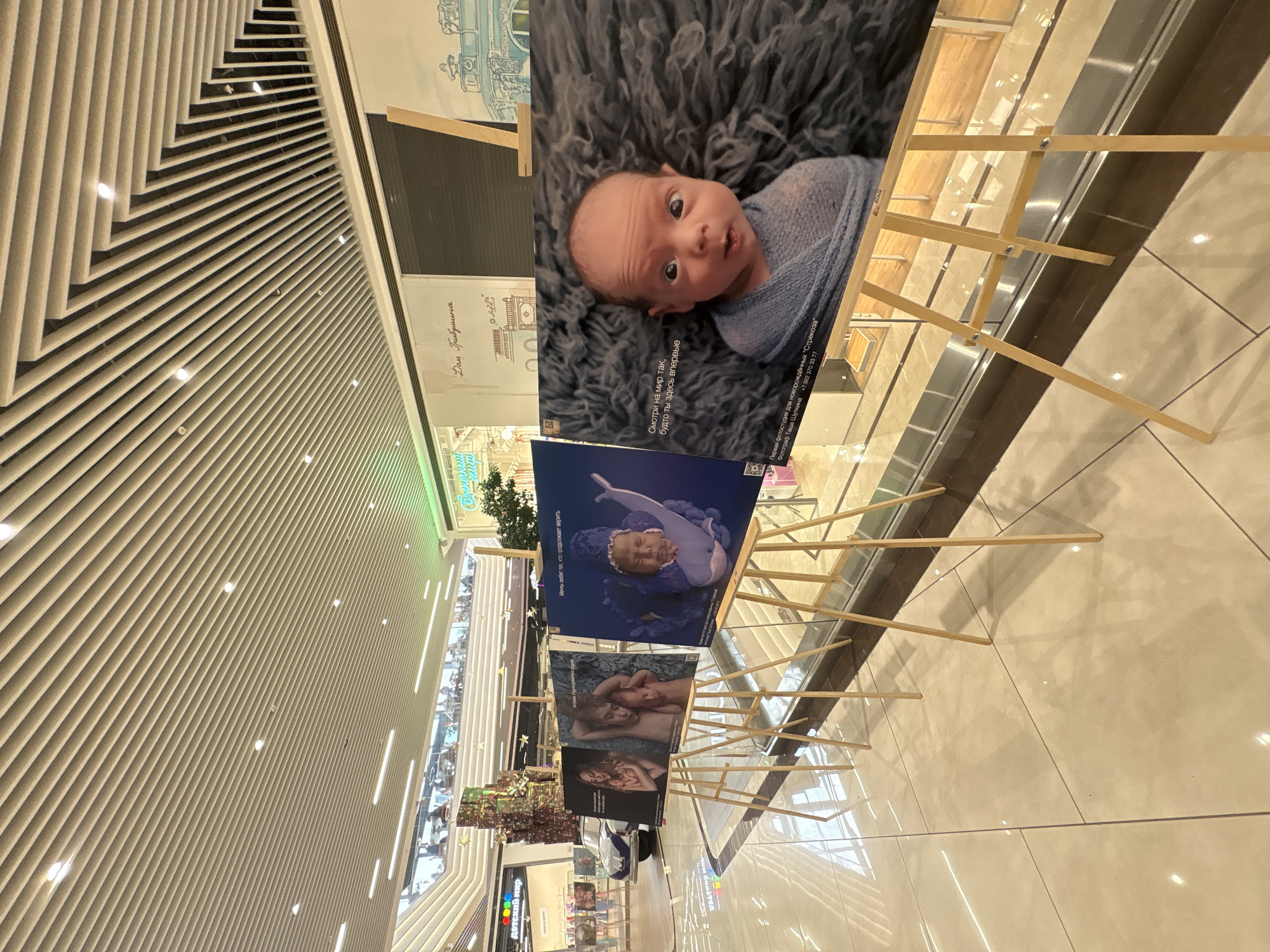 This image depicts an indoor photography exhibition set up in a multi-level shopping mall in Perm, Russia. In the foreground, a series of large photographic prints, primarily portraits of babies and young children, are displayed on wooden easels arranged in a line. The most prominent print on the right features a wide-eyed baby wrapped in a blue blanket, lying on a grey furry surface. Russian text on this print translates to "Look at the world as if you are here for the first time," and identifies the studio as "First photo studio for newborns 'Strekoza'," with the photographer "Tasha Shupina" and a contact number. Other prints show a baby dressed as a whale, a mother holding a baby, and two children together.
In the midground, to the left of the exhibition, a light blue classic-style car, adorned with greenery and flowers, is parked near a storefront. Beyond the car, a "Детский мир" (Detsky Mir) store, a well-known children's goods retailer, is visible with its distinctive logo. Another sign in the midground reads "Дом Грибушина" (Dom Gribushina), referring to a prominent location or establishment. Shoppers are visible on upper levels of the mall, which features curved architectural elements and glass railings.
The environment is brightly illuminated by various light sources, including recessed lights and long strip lights integrated into the slatted, geometrically patterned ceiling. The floor consists of large, polished, light-colored tiles, reflecting the overhead lighting and elements of the scene. The background shows more of the expansive mall interior with additional retail spaces and architectural details.