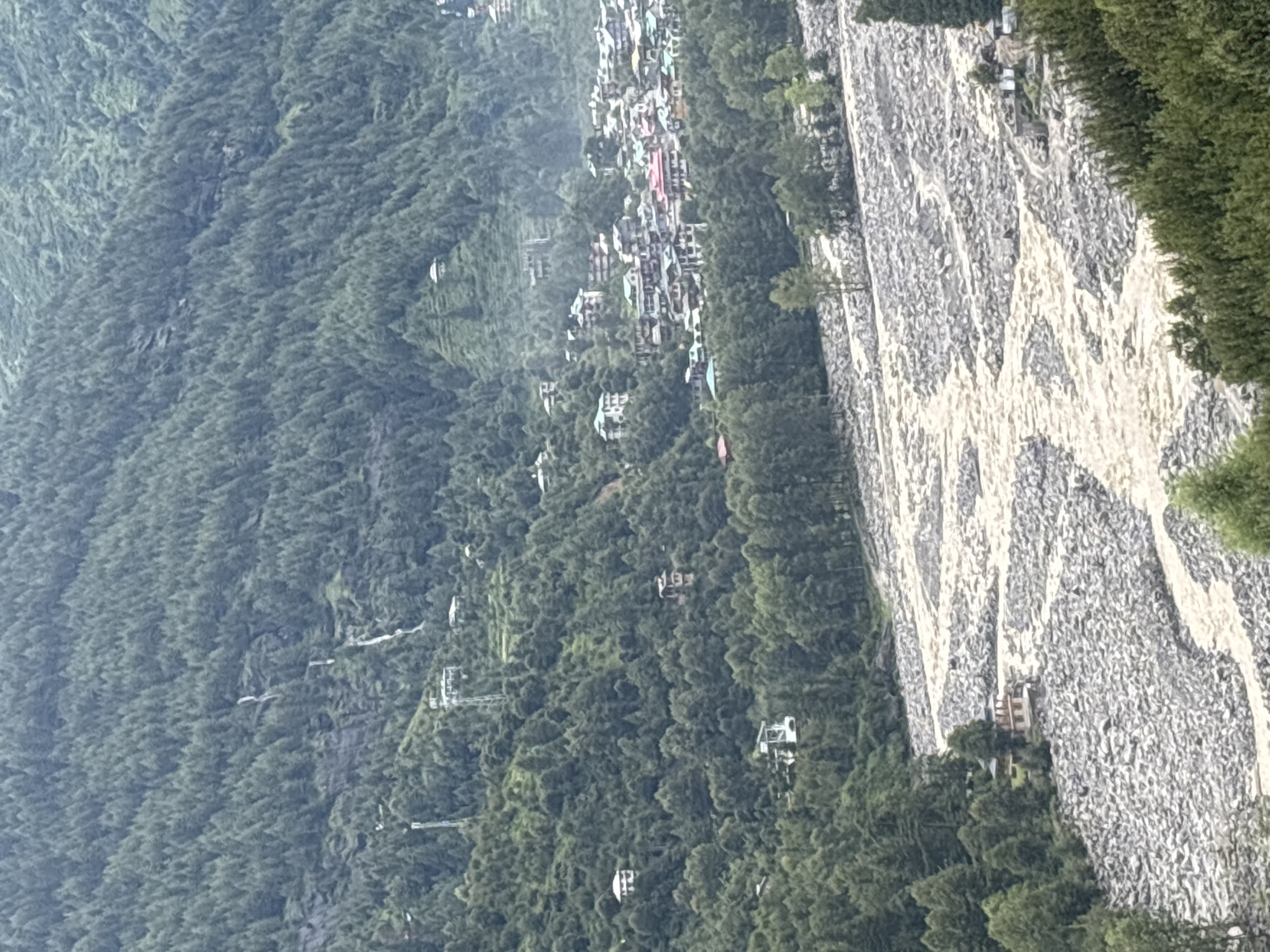 A wide, panoramic shot captures a mountainous landscape near Vashisht, India, dominated by dense evergreen forests that cover steep slopes. A muddy, fast-flowing river snakes through the lower portion of the frame, its banks lined with large, grey rocks and smaller trees. A small village, nestled amongst the trees on a hillside, features buildings with colorful roofs and balconies. Higher up the mountain, a few isolated houses are visible, some with white walls and tiled roofs. In the distance, cascading waterfalls streak down the rocky, forested mountainside. A cable car traverses the forest, connecting two points on the mountains. The overall scene depicts a rugged, natural environment interspersed with human habitation.