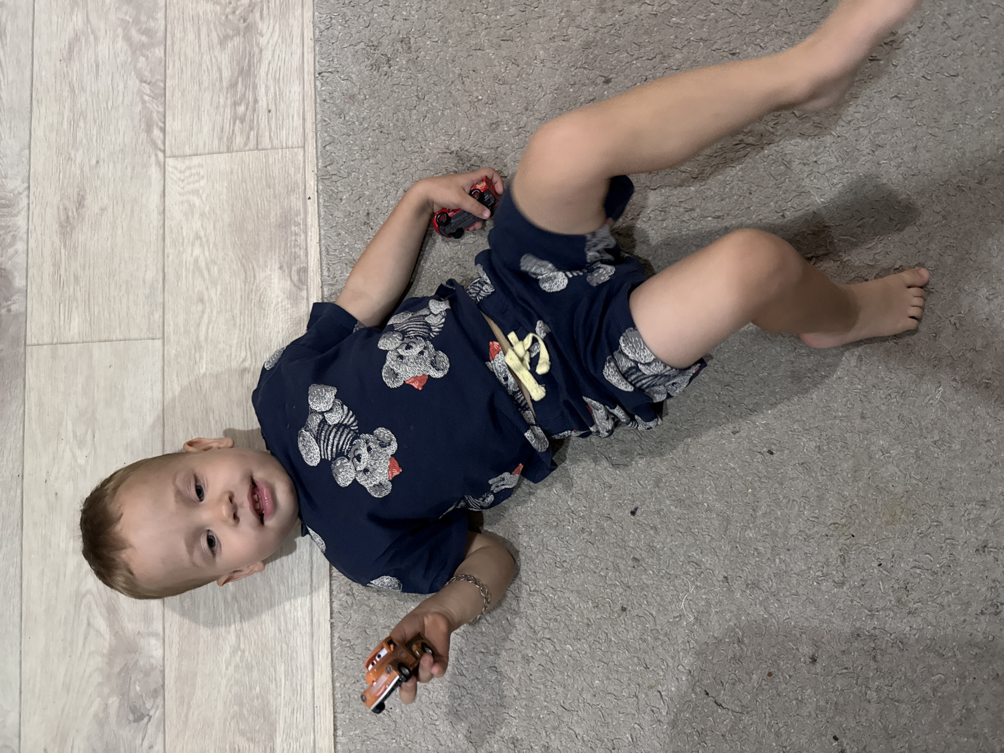 A young boy, estimated to be between 2 and 3 years old, lies on a carpeted floor next to a wooden surface, possibly a floor or wall. The boy is wearing a dark blue t-shirt and shorts with a teddy bear pattern. He is holding two small toy cars, one in each hand. The left hand holds a red and orange toy car, and the right hand holds a red toy car. The boy's body is angled, and his legs are bent. The background consists of the carpet and the wooden surface. The lighting appears to be natural, casting a shadow of the boy on the wooden surface. The overall scene is indoors and appears to be in a residential setting in the city of Rusanovo, Russia.