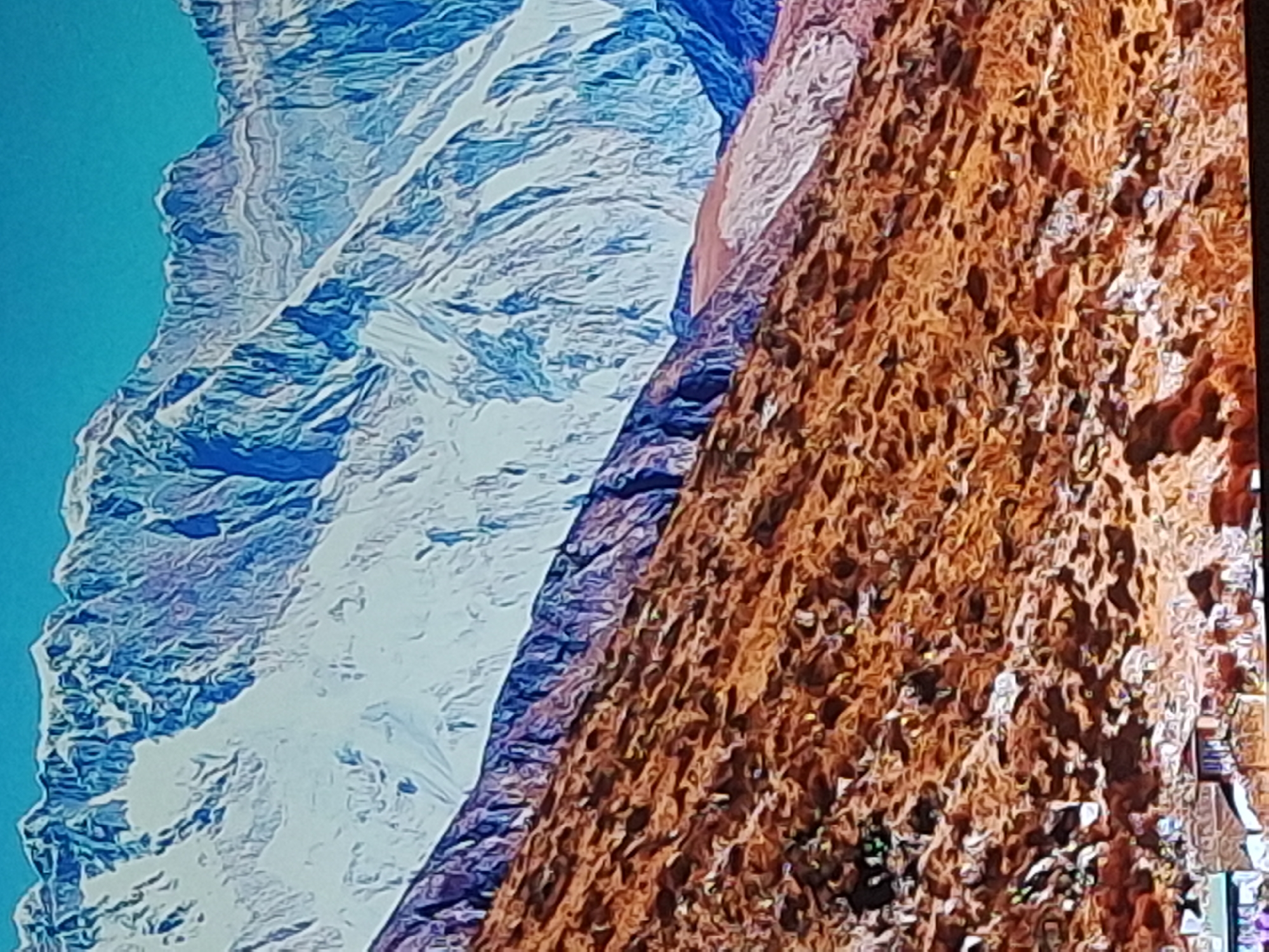 The image is a landscape photograph featuring a mountain range under a bright blue sky. The lower portion of the mountainside is covered in reddish-brown, dry vegetation, appearing rugged and textured. Above this, there is a band of darker, rocky terrain, and then the upper parts of the mountains are covered in snow and ice, with visible slopes, ridges, and shadows that highlight their dramatic contours. The snow-covered peaks are a stark white with striking blue shadows, suggesting sunlight from a high angle. A small village with buildings can be seen at the very bottom left of the image, nestled against the base of the mountain. The overall scene conveys a sense of vastness and natural beauty, with a clear delineation between the arid lower slopes and the snow-capped summits. There is no discernible location in the city of Izmail, Ukraine; the image depicts a mountainous region.