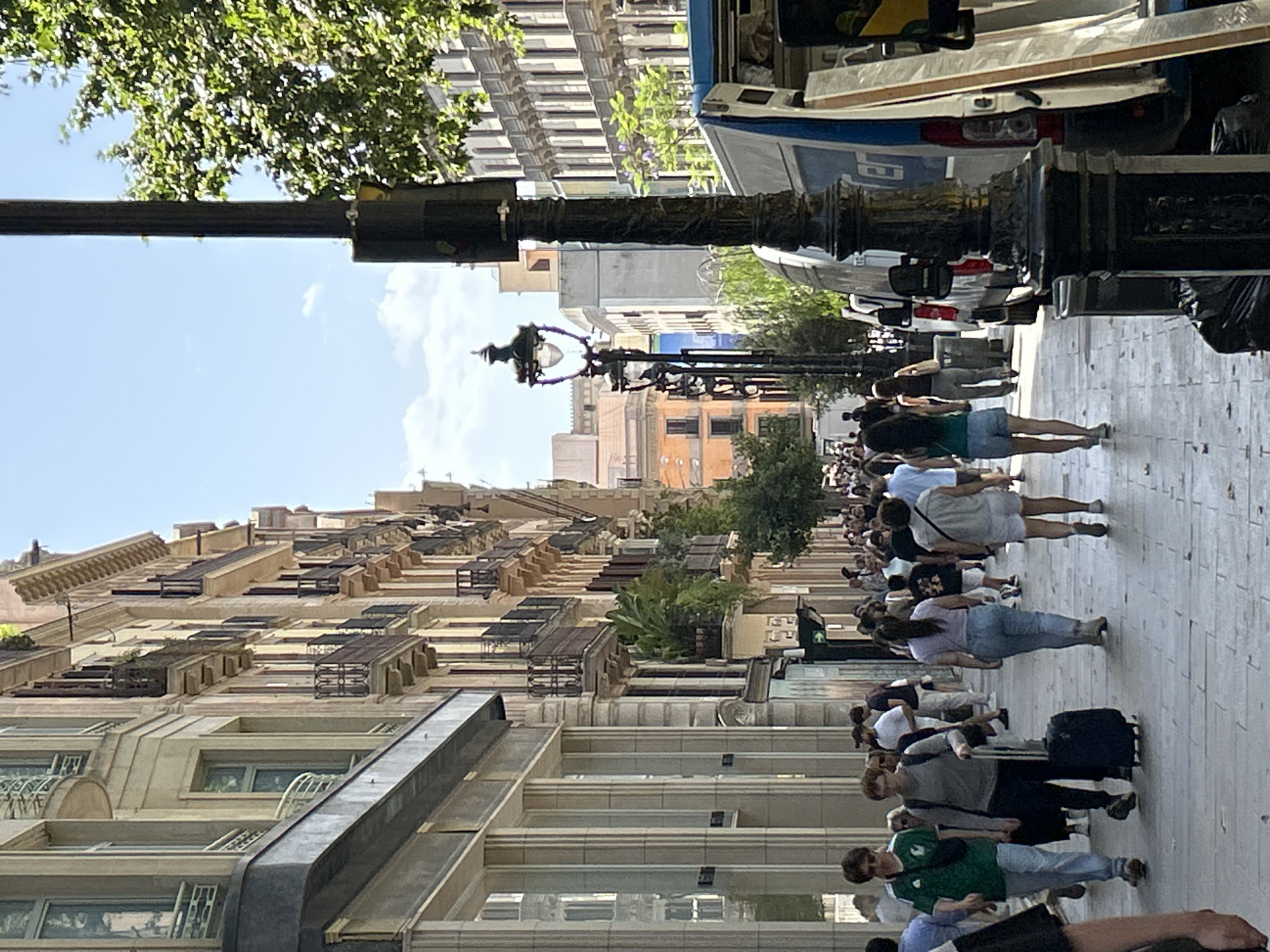 The image captures a street scene in Barcelona, Spain, on a bright, sunny day. The perspective is from a pedestrian's eye level, looking down a wide, tree-lined avenue. On the left, grand, ornate buildings with multiple stories, balconies, and large windows dominate the view. The architecture suggests a historic European city. On the right side of the street, more buildings are visible, including some with vibrant orange facades. A dark lamppost with intricate detailing stands prominently in the foreground on the right, and further down the street, another similar lamppost is visible.
A moderate number of people are walking along the sidewalk. They are dressed in casual summer clothing, suggesting warm weather. Some individuals are seen with luggage, indicating travel. People are walking in both directions, some in small groups, others individually. The sidewalk is paved with cobblestones or similar material.
In the background on the right, a white van with blue lettering is parked, and a portion of what appears to be a delivery truck or trailer is visible next to it, with some long, flat items leaning against it. Green trees with lush foliage line the right side of the street, providing shade. The sky is clear blue with a few wispy clouds. The overall impression is a bustling urban environment, likely a popular shopping or tourist area in Barcelona.