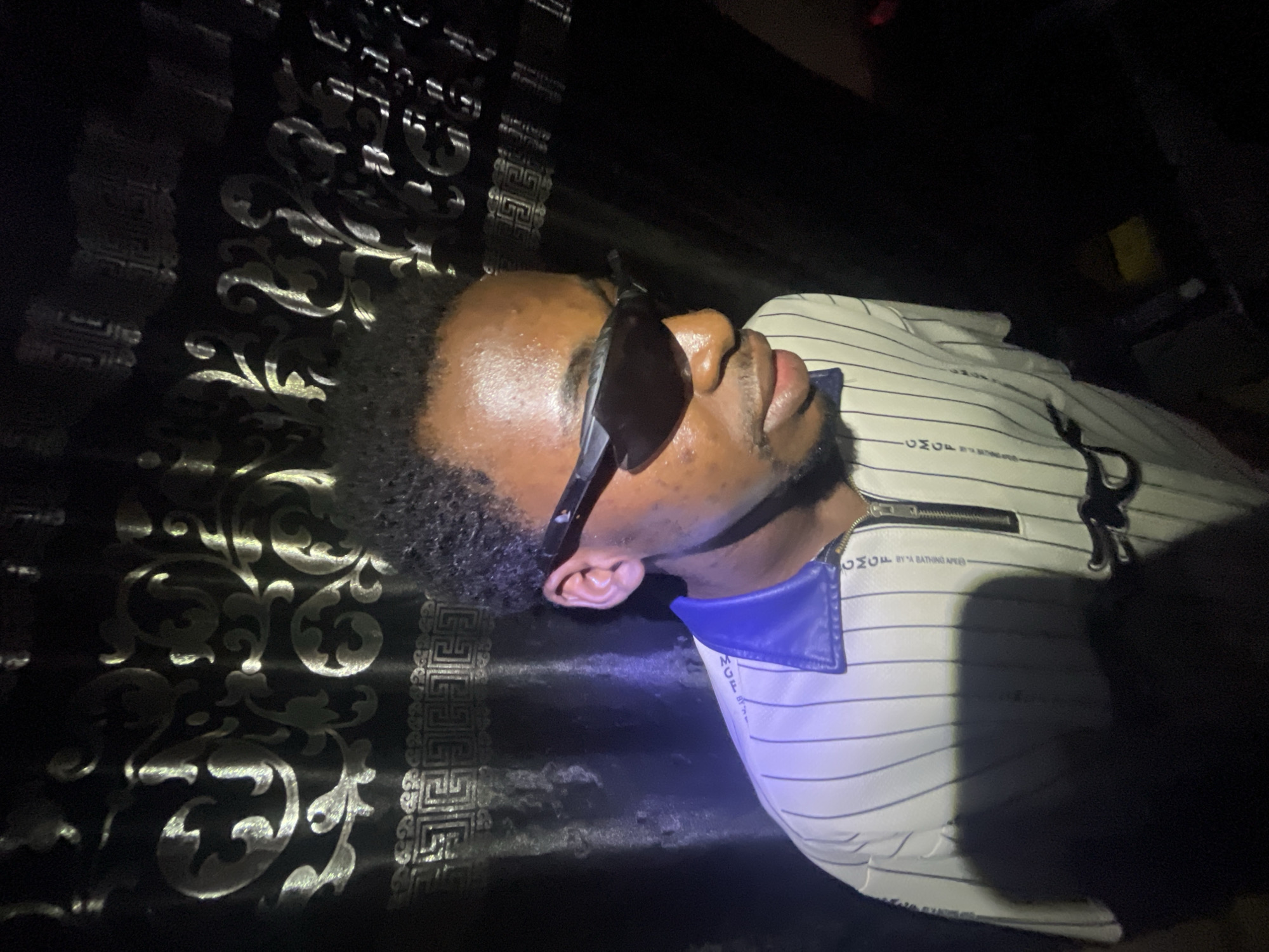 A person wearing sunglasses and a striped shirt with a blue collar is lying on what appears to be a patterned surface. The location of the image is indeterminate, as there is no verifiable information to conclude it is in Lokoja, Nigeria. The environment appears to be dimly lit.