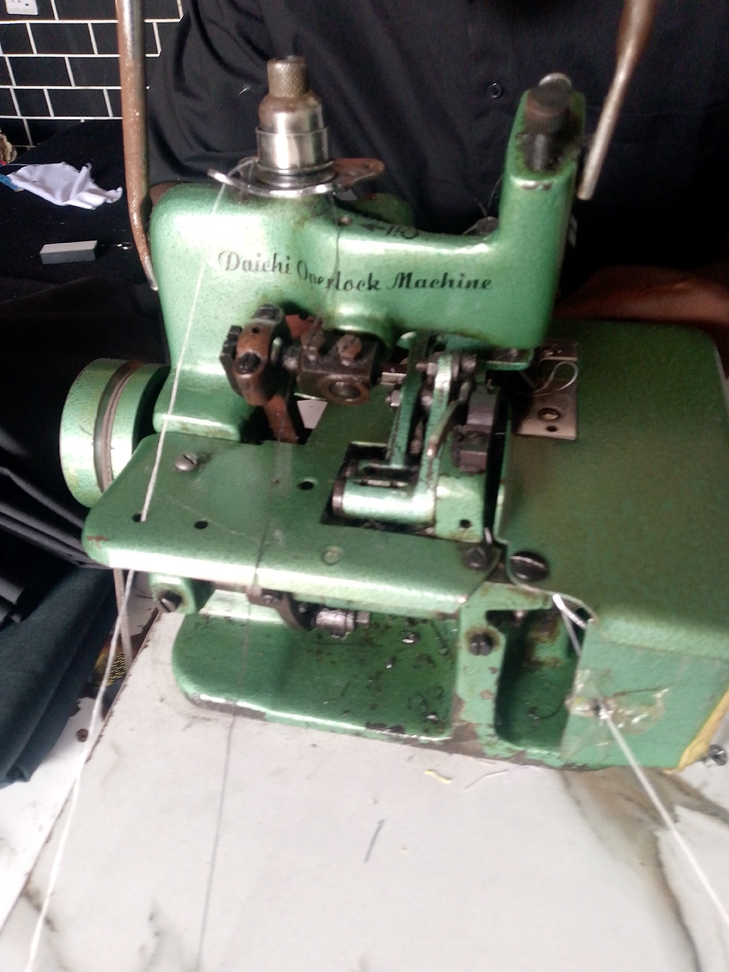 In Jauro Yinu, Nigeria, a Daichi Overlock Machine, painted green, sits on a white surface. The machine is a sewing machine, characterized by its multiple needles and threads. It is operational, with white thread running through it. The machine is partially obscured by a person wearing a black shirt. The background is blurred, but suggests a domestic or workshop environment with dark cabinetry and what appears to be a brick wall. The overall scene depicts a functional sewing machine in a modest setting.