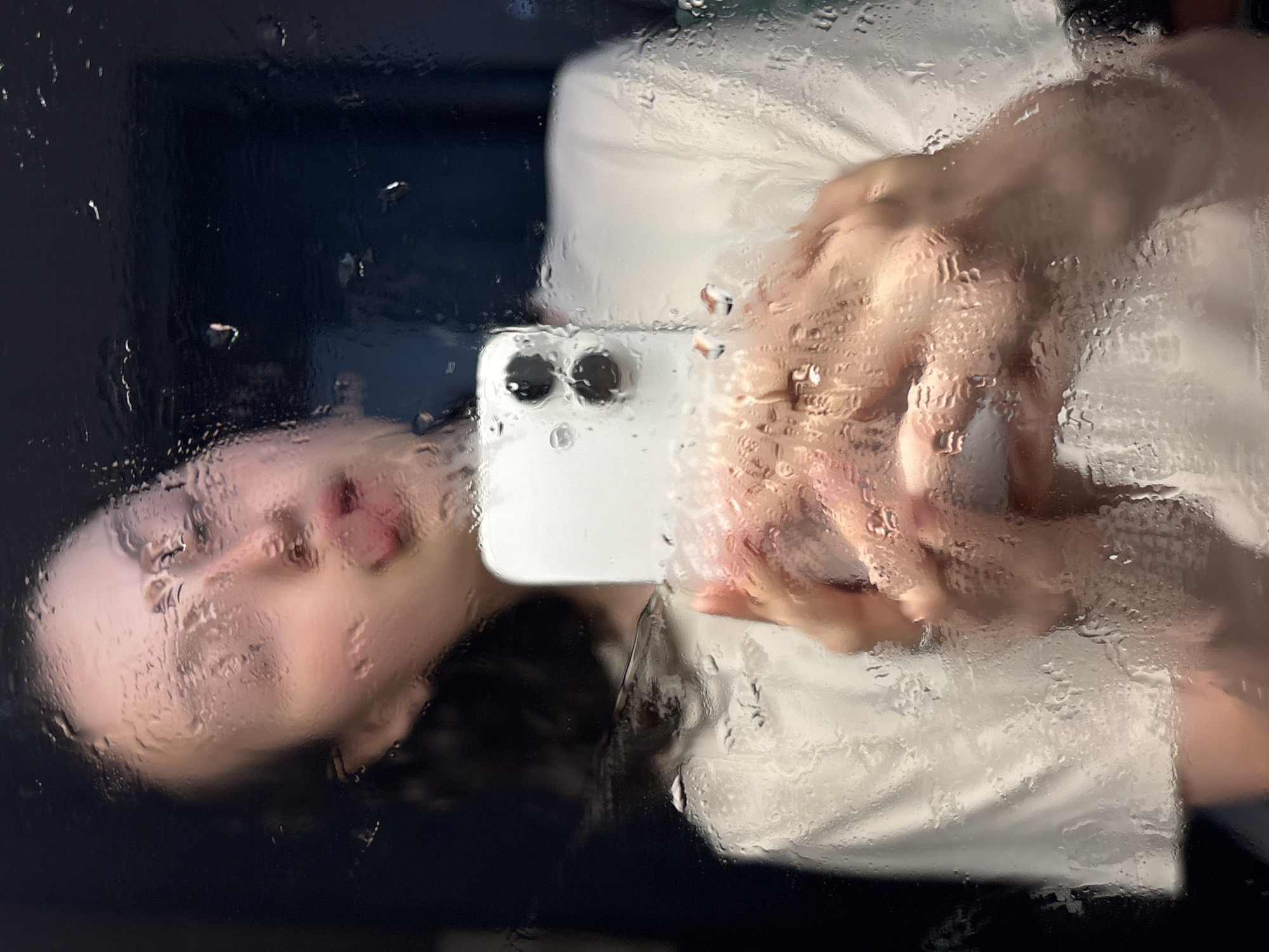 A young woman with dark hair is taking a selfie in a mirror covered in water droplets. She is wearing a white t-shirt and is holding a white smartphone up to the mirror. Her eyes are closed, and her tongue is sticking out. The background is dark and out of focus. The location is not specified in Tikhiye Zori, Russia.