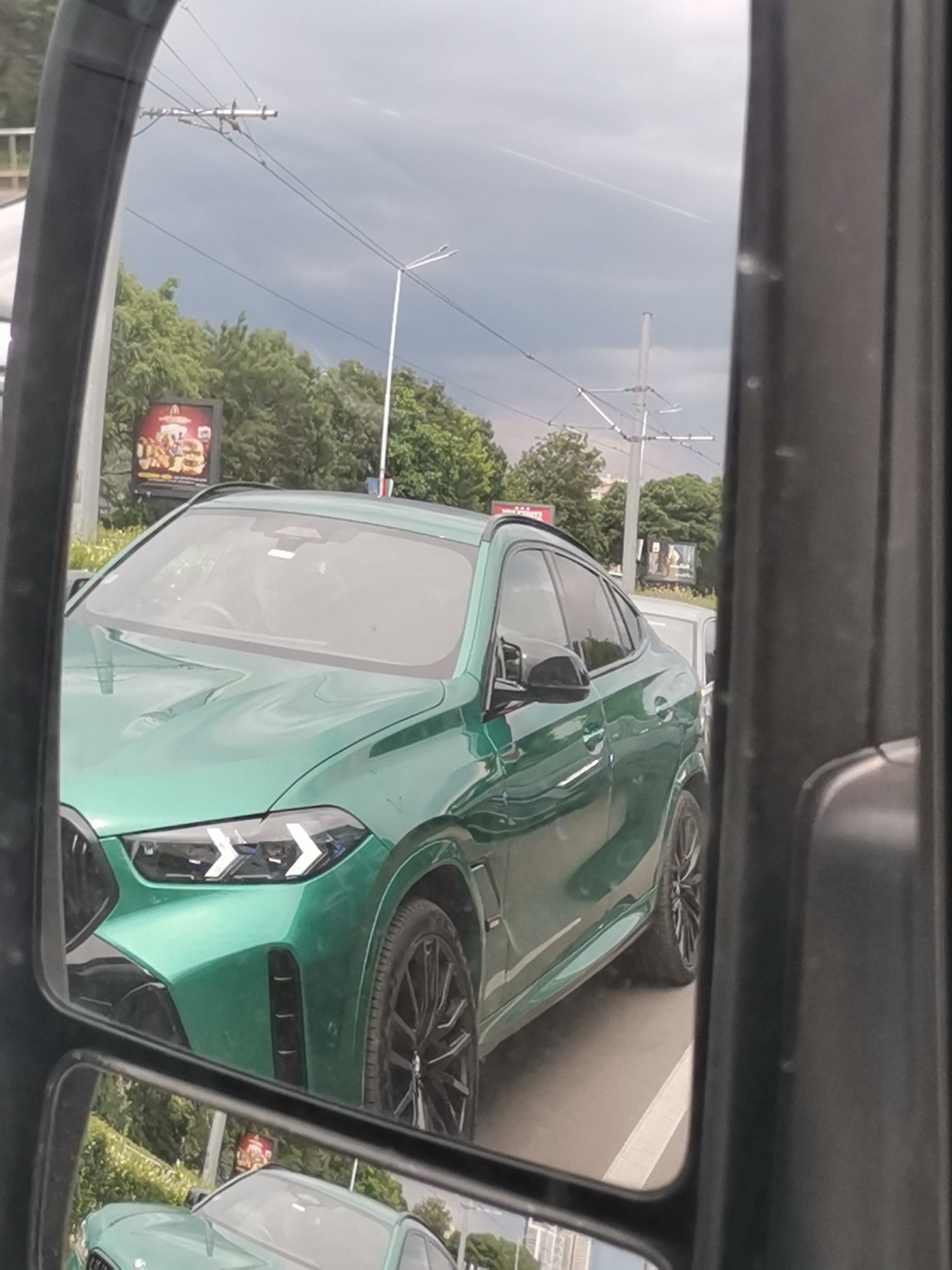 The image is a reflection in a car's side mirror, showing a green BMW X6 M. The car is stationary in traffic. In the background, there are trees, utility poles with overhead wires, a McDonald's billboard, and a cloudy sky. The reflection also shows other vehicles and buildings. The image was taken in Sofia, Bulgaria.