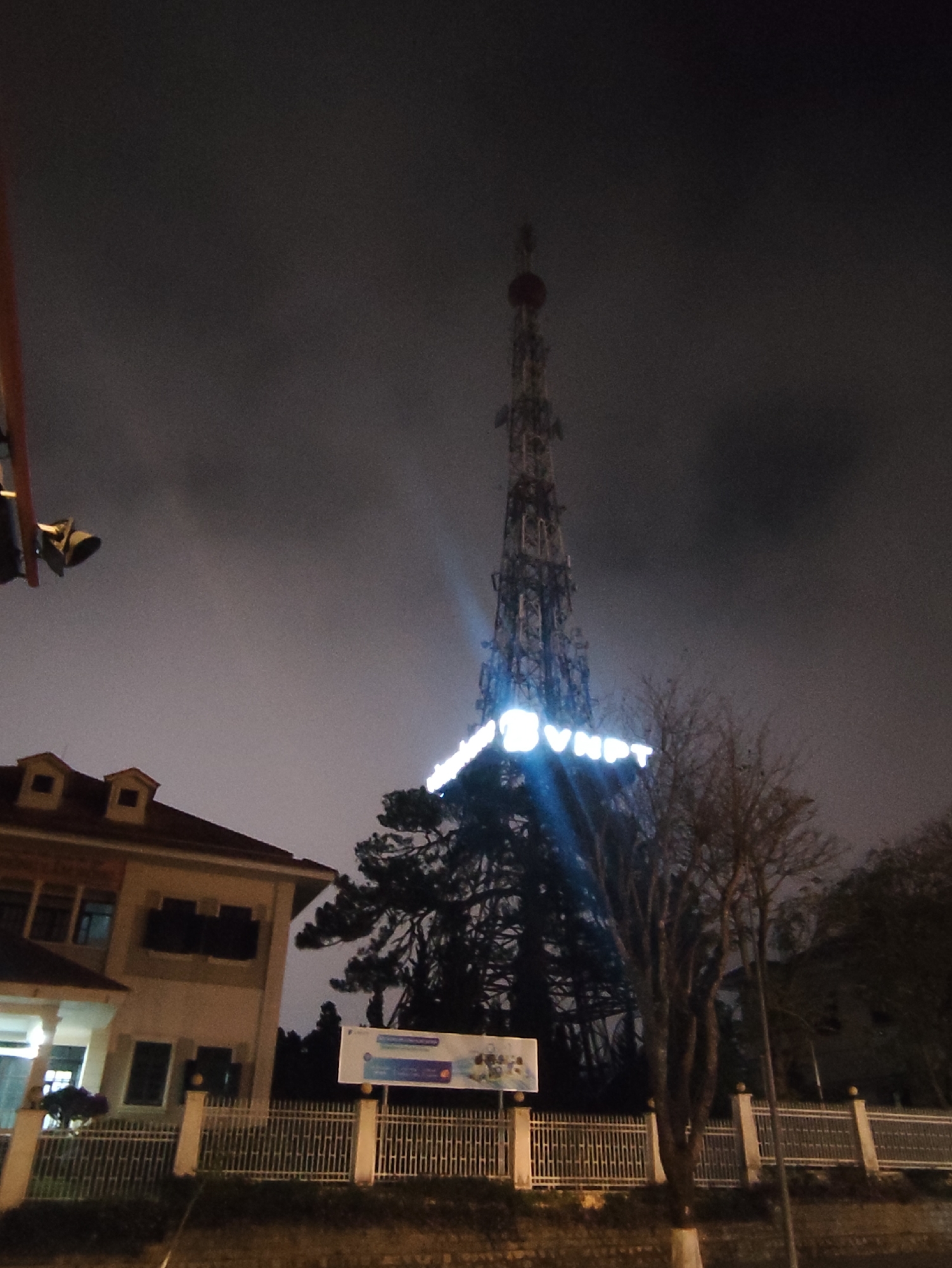 At night in Da Lat, Vietnam, a tall VNPT telecommunications tower is visible against a dark, cloudy sky. The VNPT logo is illuminated in white. The tower is situated behind a building with a red-tiled roof and a fence. A sign is visible near the fence. Bare trees are also visible in the scene.