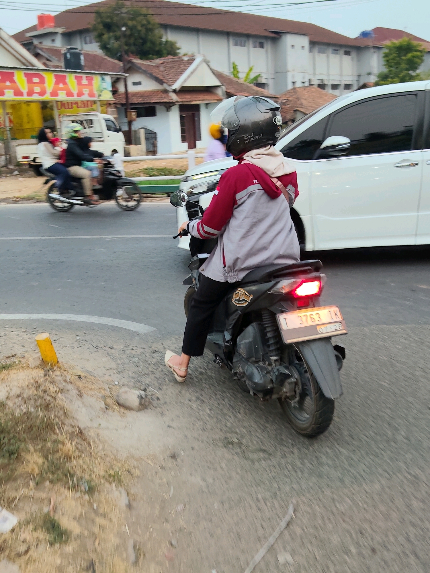 A person on a dark-colored scooter is captured from behind, riding on a paved road. They are wearing a gray jacket with red accents, black pants, and white sandals. A dark helmet with a clear visor adorns their head. The scooter's license plate reads "T 3763 IK". In the background, a white van is parked, and beyond that, buildings with red tile roofs and white walls are visible. A yellow pole stands on the side of the road. The scene appears to be in an urban setting in Purwakarta, Indonesia, possibly during the day.