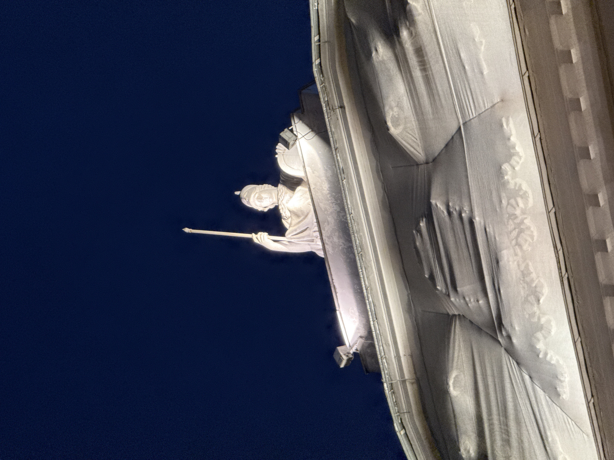 The image shows a statue on the roof of a building at night. The statue is white and appears to be of a person holding a staff. The building is covered in a white tarp, and there are lights illuminating the statue and the tarp. The sky is dark blue. The building is the Former Russian State Academy of Theatre Arts, located in the city of Saint Petersburg, Russia.