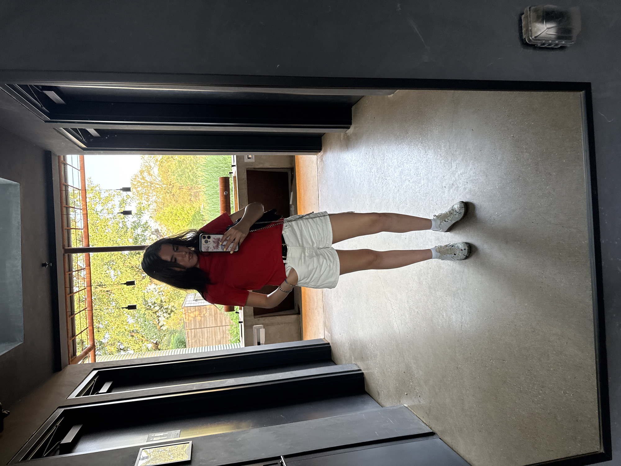 The image shows a young woman taking a selfie in a mirror. The background reveals an indoor space with an exterior view of trees and a building, suggesting the photo was taken in a building with an open view. The location is identified as Sunset Valley, United States of America. The woman is wearing a red shirt, white shorts, and white sneakers. She is holding a smartphone in her hand. The mirror is framed in black. A light fixture is visible on the wall near the mirror. The floor is a smooth, light-colored material. The indoor space has a modern, architectural design with metal beams and window frames.