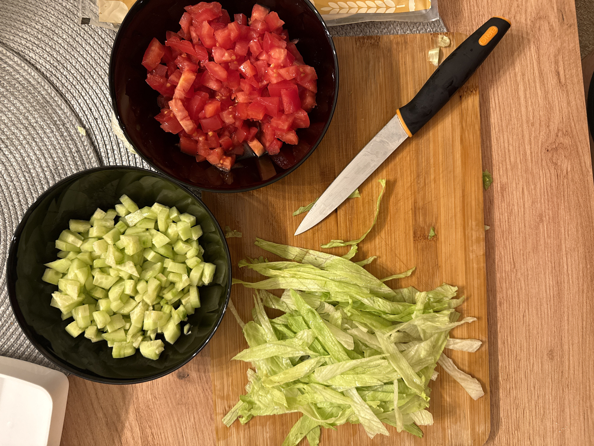 An overhead shot captures a scene of food preparation on a wooden surface, likely a kitchen counter or dining table. The primary subjects are three bowls of chopped vegetables and a knife on a cutting board.
In the upper left, a black bowl contains a generous portion of diced red tomatoes, uniformly cut. Below it, another black bowl holds an equally large quantity of diced light green cucumbers. To the right of the cucumber bowl and below the tomato bowl, a pile of thinly shredded light green lettuce is spread across a light-colored wooden cutting board.
A chef's knife or utility knife rests diagonally on the cutting board, its silver blade pointing towards the bottom left. The knife has a black handle with an orange trim, and the brand name "BRIO" is visible in orange text on the handle. Small remnants of lettuce leaves are scattered around the knife and on the cutting board, indicating recent chopping activity.
Part of a grey, woven placemat is visible under the bowls on the left side of the frame. In the top right corner, a portion of a light-colored paper or cardboard package, possibly food packaging, is partially visible. The lighting is artificial, suggesting an indoor setting, potentially during the evening. No people are visible. The overall scene depicts ingredients being prepared for a fresh meal or salad.