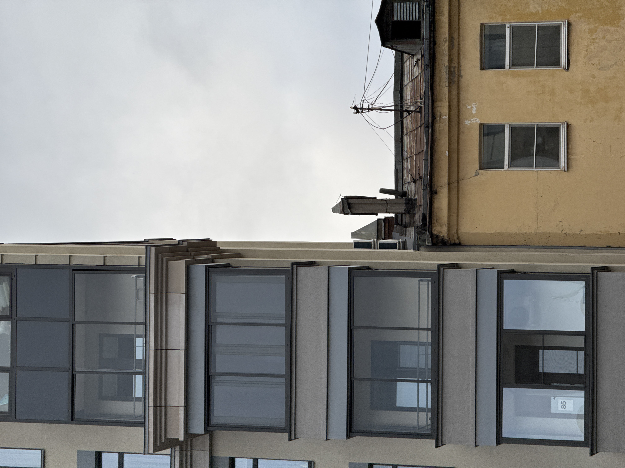 The image shows an urban scene in Saint Petersburg, Russia, focusing on the facades of two adjacent buildings. The building on the left is a modern, multi-story structure with a light beige exterior and dark-framed windows, some of which appear to be enclosed balconies. The windows are large and rectangular, arranged in horizontal bands across the building. One window on a lower floor has a white sign with the number "85" visible.
To the right of the modern building is an older, lower building with a weathered yellow stucco facade. This building has a sloping, rusty metal roof, two visible windows with white frames, and several chimneys and ventilation structures on the roofline. A complex network of wires and a utility pole are also present above this roof. The sky is overcast and a dull grey. The juxtaposition of the two buildings highlights the architectural contrast between old and new within the city. No people are visible in the image.