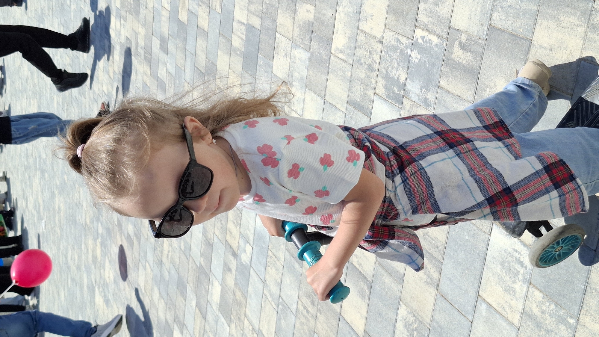 A young girl rides a scooter outdoors in Unknown, Unknown. She is wearing sunglasses, a white shirt with pink flower patterns, a plaid shirt tied around her waist, and jeans. She holds the teal handlebars of the scooter.
To the left, portions of several people are visible, including legs in jeans and black pants. A pink balloon is also present.
The ground is paved with grey and white rectangular blocks.