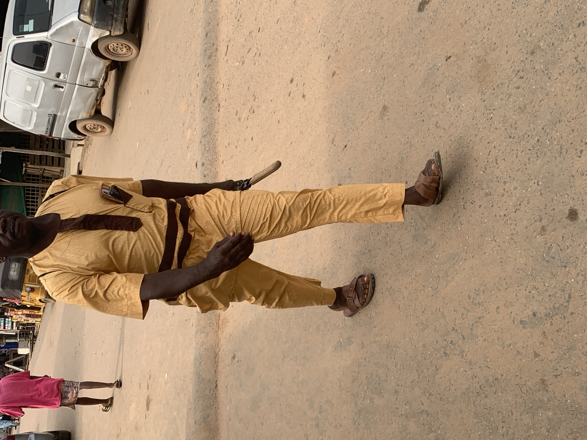 The image shows a man walking on a dusty road in Shagamu, Nigeria. He is wearing a traditional yellow garment with a dark brown sash and matching pants. He has brown sandals on his feet and a small object, possibly a phone, tucked into his chest pocket. He appears to be walking away from the camera, and his right hand is extended slightly. In the background, a silver van and a yellow tricycle are visible, along with some market stalls. The road surface is asphalt with a significant amount of dust and debris. Another person in a pink shirt and patterned skirt is walking away in the distance on the left side of the frame.