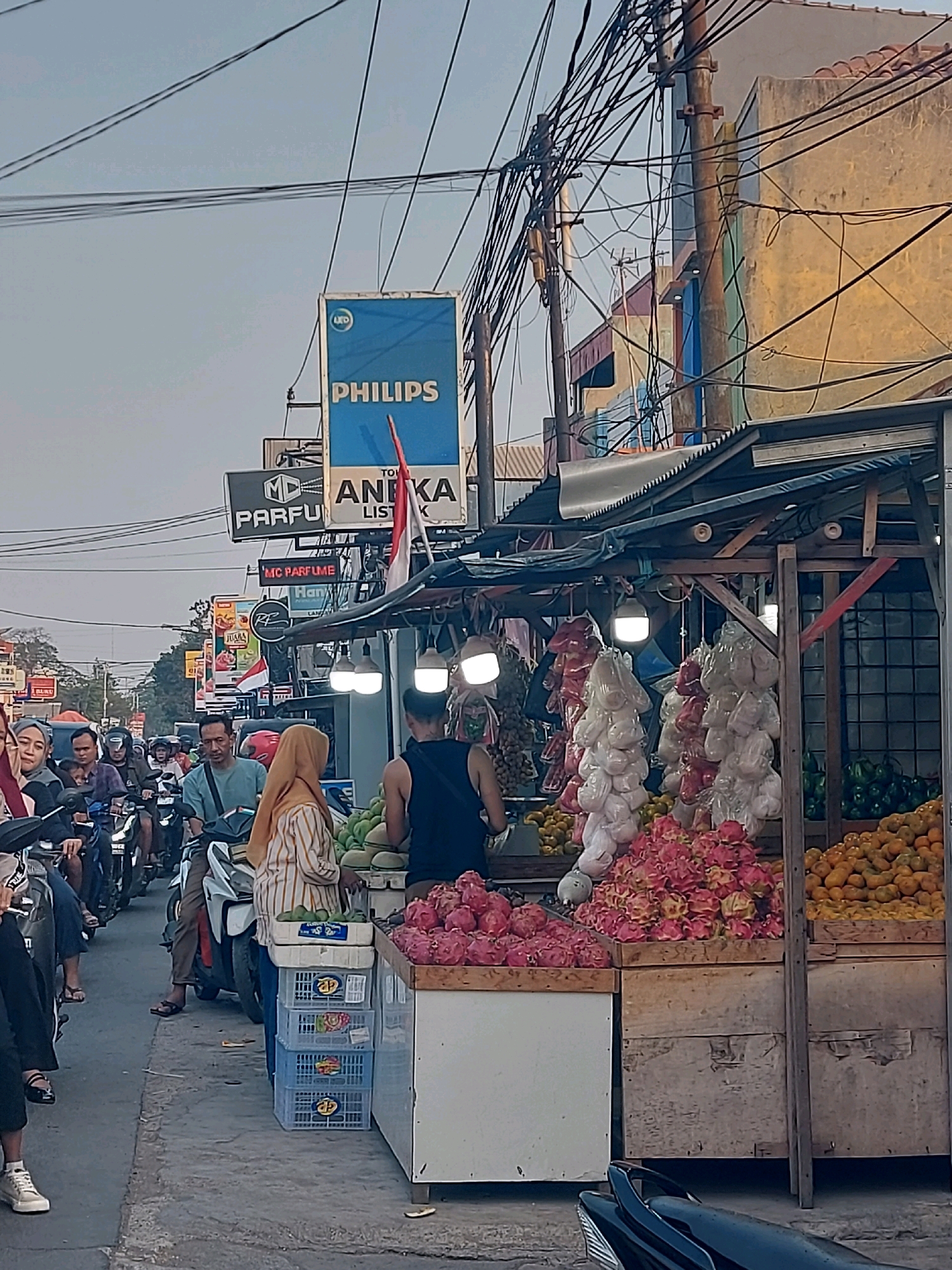 This is an eye-level, medium shot of a street scene in Karawang, Indonesia, featuring a fruit stand on the right and a traffic jam on the left. The sky is overcast. A wooden fruit stand, partially covered by a tarp, displays an assortment of fruits, including dragon fruit, oranges, and what appear to be melons. Several plastic-wrapped bunches of what seem to be garlic or onions hang from the stand. Two women are visible at the fruit stand; one, wearing a yellow hijab and striped shirt, appears to be attending to the fruits, while the other, in a black tank top, stands to her right. In the background, a "PHILIPS" sign is prominent, along with other advertisements and a dense network of electrical wires.
To the left of the fruit stand, a traffic jam of motorcycles and scooters is depicted. Several individuals are on the motorcycles, some wearing helmets. One man in the foreground, on a motorcycle, is looking to his left. The road is paved, and a sidewalk area is visible in front of the fruit stand. The overall impression is a bustling urban environment with a mix of commerce and daily life.