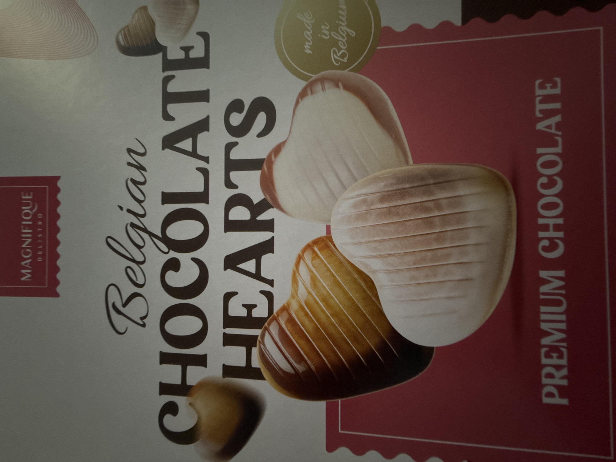 This is a close-up, angled shot of the packaging for "Belgian Chocolate Hearts." The image primarily displays a product box featuring an arrangement of heart-shaped chocolates.
The packaging has a split design: the left side is a light gray or off-white, and the right side is a solid reddish-pink color with a scalloped border separating the two sections. Several heart-shaped chocolates are depicted on the reddish-pink background. These chocolates vary in color, showing milk chocolate, white chocolate, and some with a marbled or two-tone appearance, combining both milk and white chocolate. Each chocolate heart has distinct ridges or grooves on its surface. A smaller, darker chocolate heart is partially visible at the bottom left, extending onto the light background.
Visible text on the packaging includes:
- Prominently in the center, "Belgian CHOCOLATE HEARTS." "Belgian" is written in an elegant script font, while "CHOCOLATE HEARTS" is in a bold serif font.
- In the top left corner, within a red rectangular label with a decorative edge, the word "MAGNIFIQUE" is visible, with "DELISTRO" below it.
- In the upper right section, a circular label with a golden border and a green interior states "made in Belgium" in a script font.
- Along the right edge of the package, vertically oriented, the words "PREMIUM CHOCOLATE" are displayed in a light-colored serif font against the reddish-pink background.
The image does not show any people or specific activities. It is a static product shot. The location context provided is Varna, Bulgaria, though this is not depicted in the image itself.