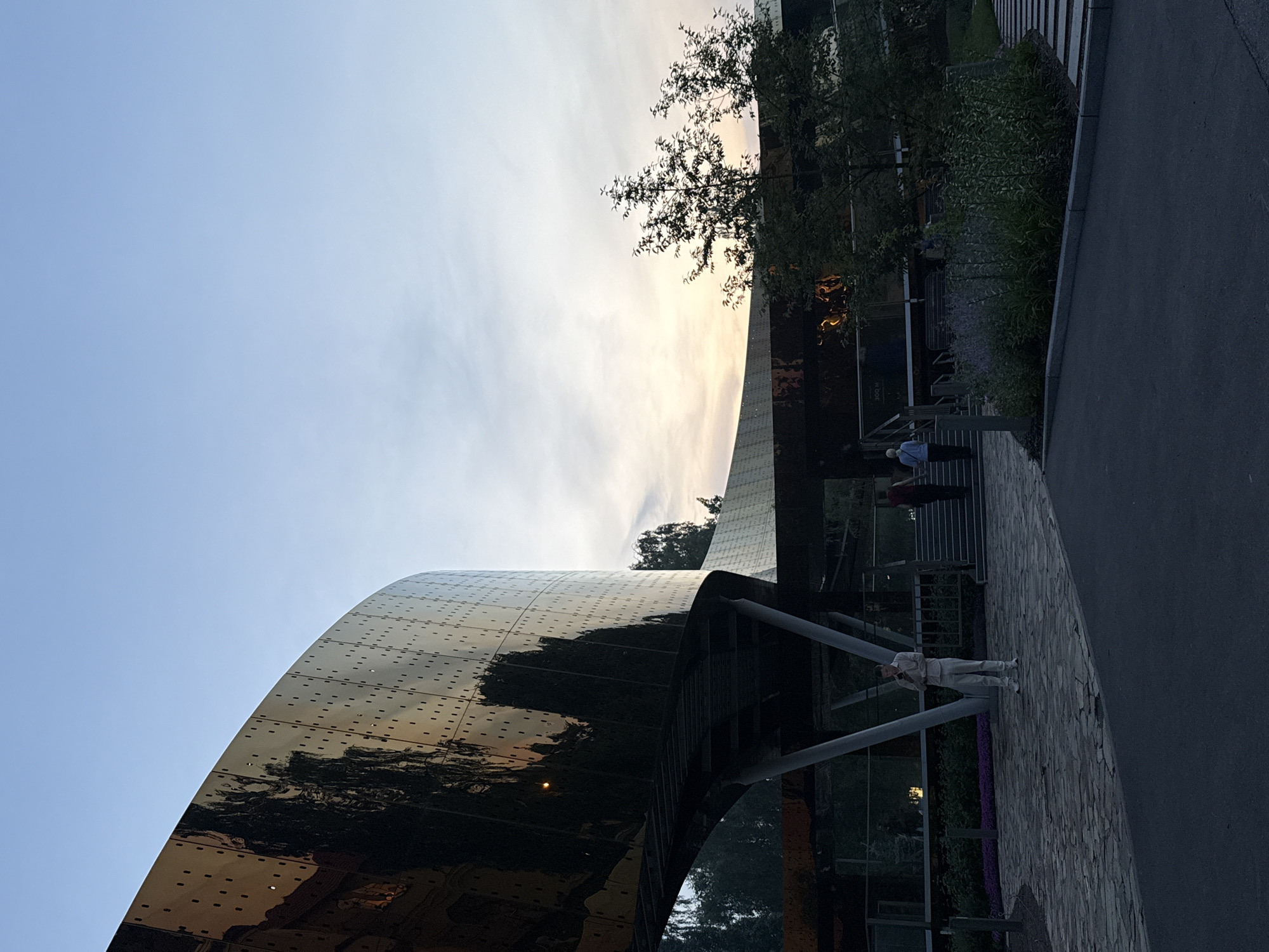 The image depicts the Zaryadye Park in Moscow, Russia, at dusk. A striking, curved, golden building, possibly a pavilion or part of a larger structure, dominates the left side of the frame. Its surface is highly reflective, mirroring the sky and surrounding trees. A walkway made of stone pavers leads towards the building. In the mid-ground, a few people are visible walking or standing, interacting with their surroundings. One person is standing in the foreground, looking at their phone. The sky is a soft gradient of blue and orange, indicating the setting sun. Lush greenery and trees are scattered throughout the park, adding to the natural ambiance. The overall atmosphere is serene and contemplative, suggesting a moment of quiet reflection in an urban oasis.
