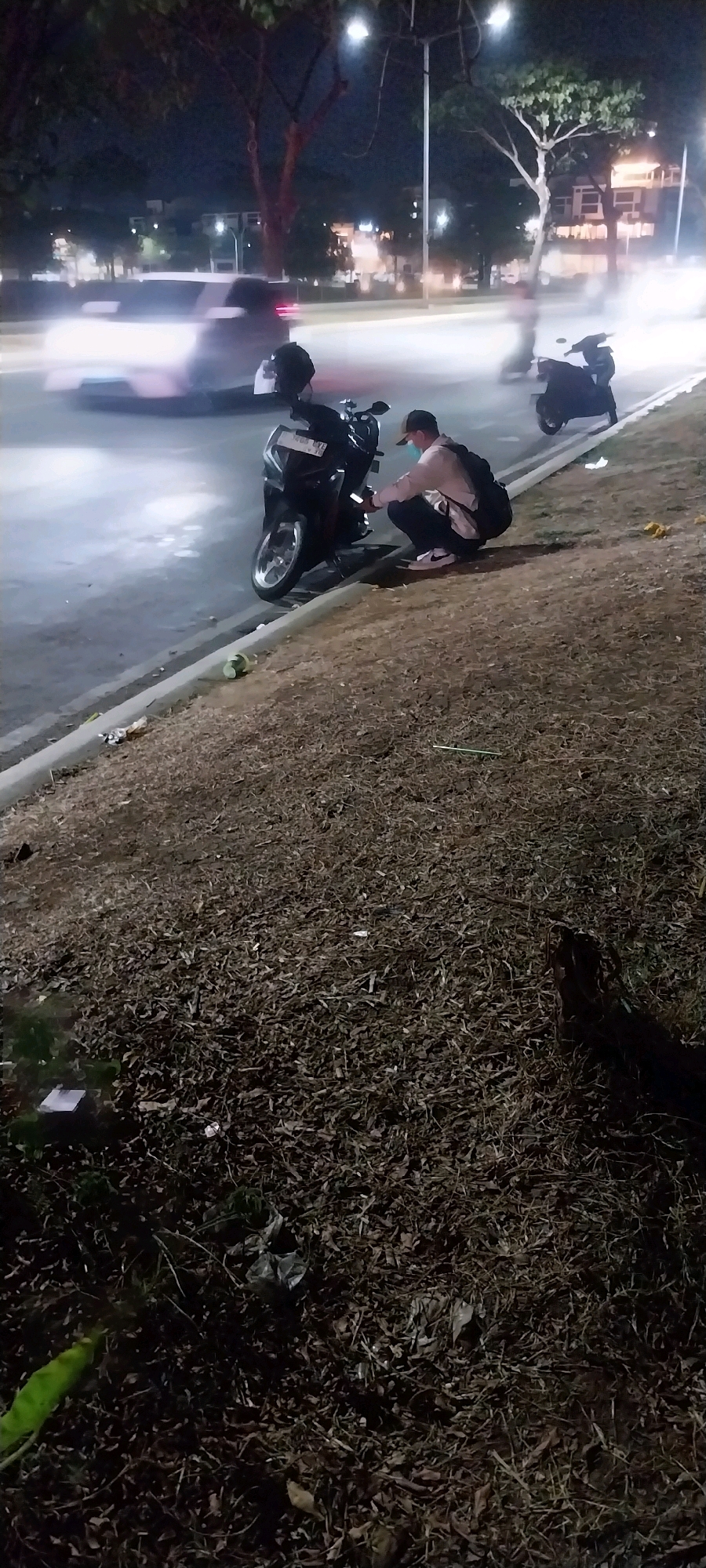A person wearing a white shirt, dark pants, and a backpack squats near a parked motorcycle on the side of a road at night in Medan, Indonesia. The person is looking at their phone, and their face is obscured by a face mask. In the background, cars are driving by, their headlights creating motion blur. The area is dimly lit by streetlights. The ground is covered in dry leaves and some trash. Another motorcycle is parked further down the road.