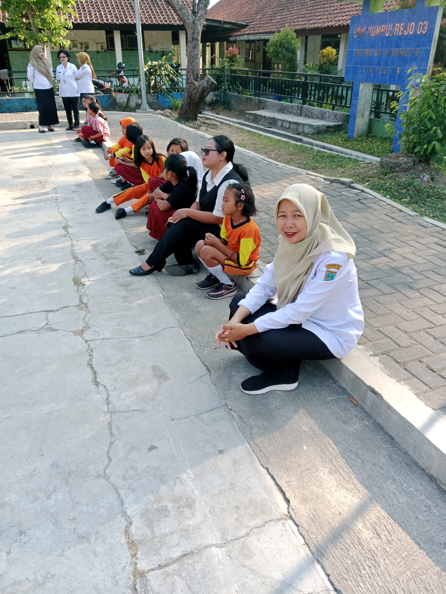 The image depicts a group of people, mostly women and young girls, sitting on a concrete path outdoors, in front of a school building. The school building has a reddish-brown tiled roof and a blue sign that reads "SD N. KUMPULREJO 03 KECAMATAN TINGKIR SALATIGA." Three adult women are visible in the foreground and background, two of whom are wearing white shirts and headscarves. One woman in the foreground is wearing a light brown headscarf, a white long-sleeved shirt with a badge on the left sleeve, and black pants, and is crouching down. Several young girls in brightly colored school uniforms (orange and red) are sitting in a line behind the adult women. The environment appears to be a schoolyard or outdoor space with a paved walkway, a grassy area, and trees. The lighting suggests it is daytime, with shadows cast by the sun.