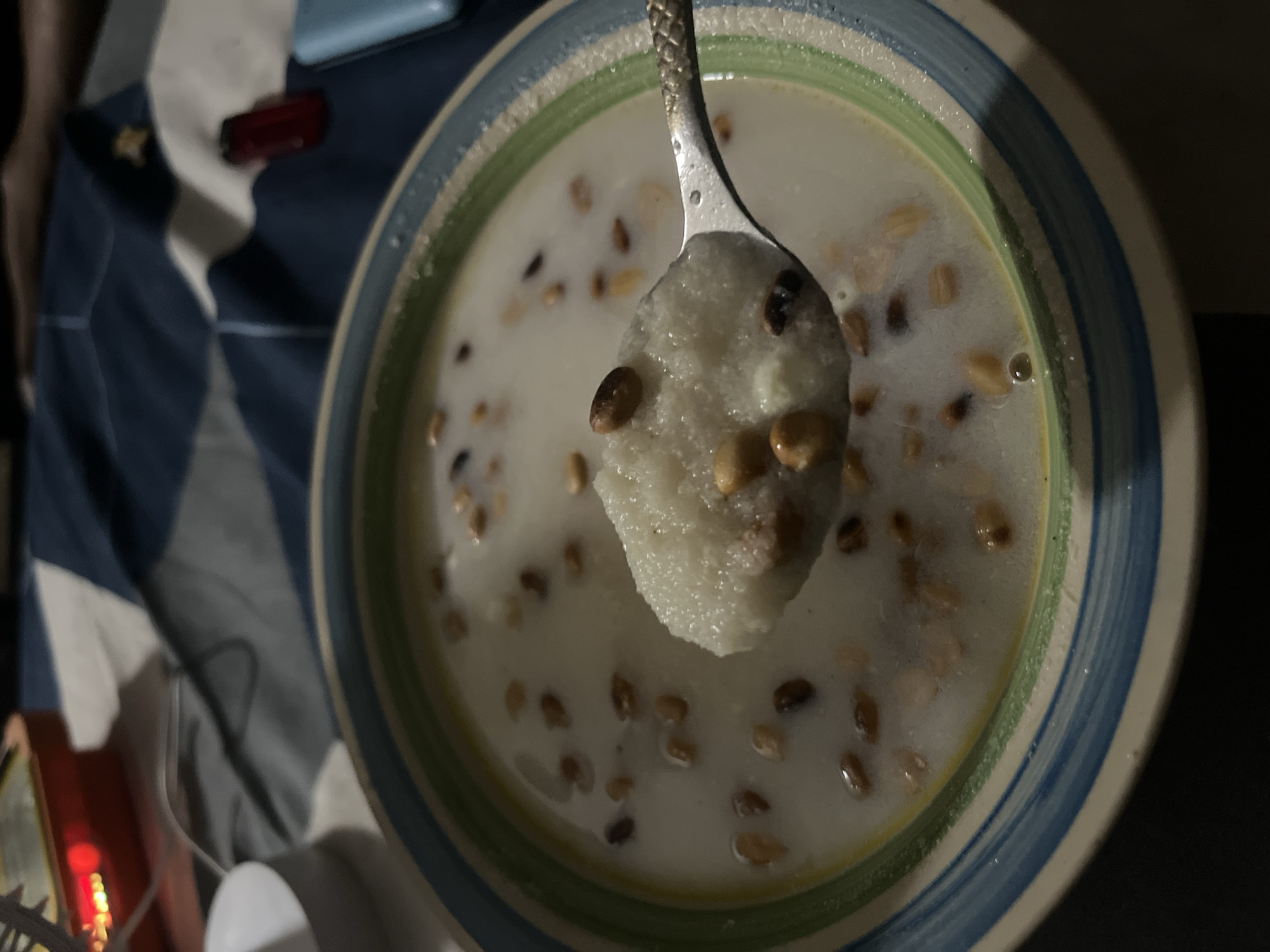 A bowl of porridge with scattered grains is held up by a spoon. The bowl has a green, blue and white rim. The location is likely a home in Lokoja, Nigeria, as implied by the prompt. Other objects present include part of a bed with blue bedding, a red object, and a white container. The scene is dimly lit. There are no people distinctly visible, although an arm is slightly visible. There are no interactions taking place in the image. The porridge seems to be the central subject of the image.