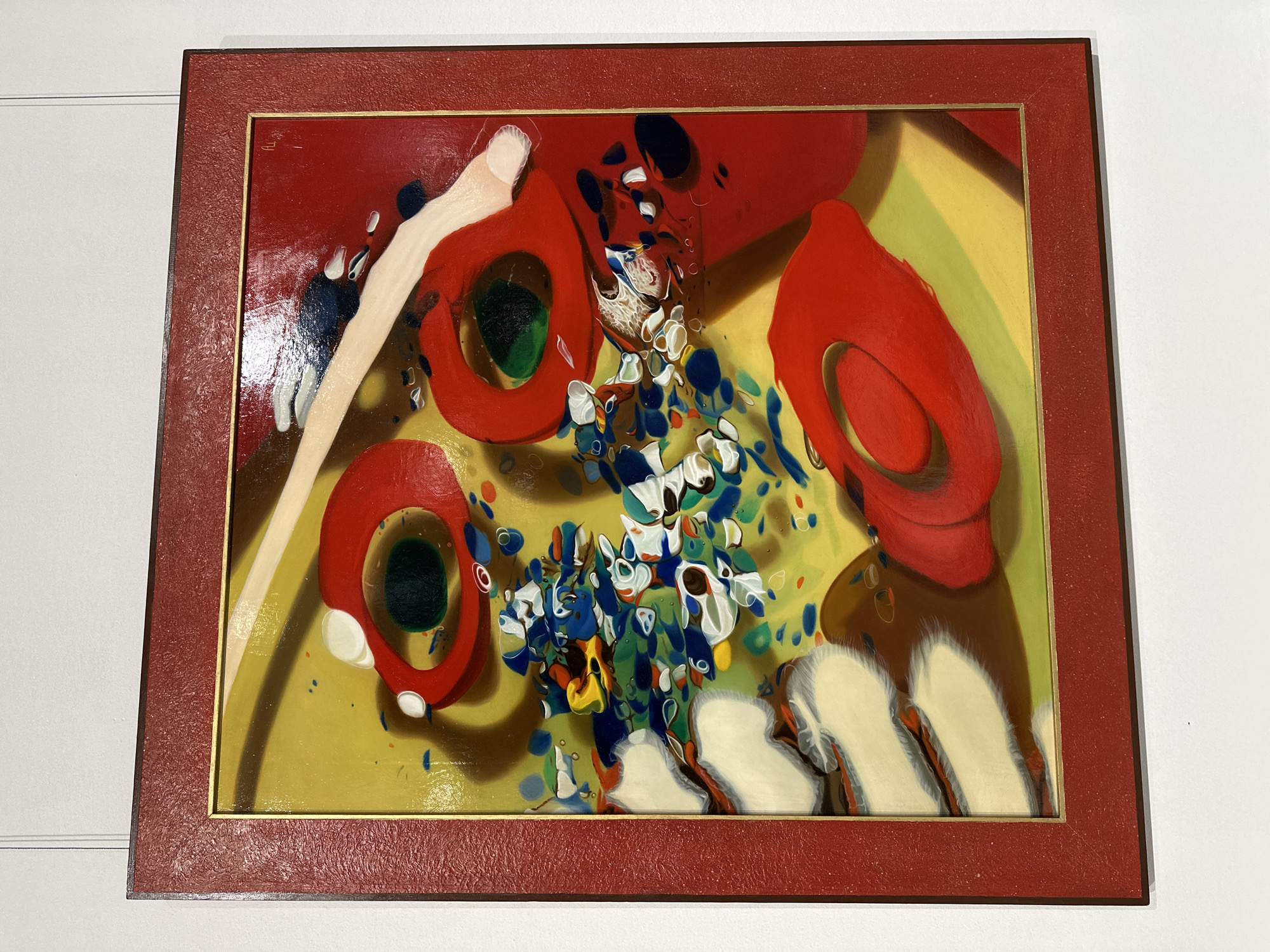 An abstract painting, enclosed in a reddish-brown textured frame with a thin gold inner border, hangs on a light-colored wall. The artwork features a vibrant and dynamic composition dominated by bold colors and organic, fluid shapes.
The painting's background is a bright yellow-green field, with areas of warmer yellow and brown creating depth. Several large, glossy red forms resembling stylized hats, bowls, or perhaps even lips, are prominent. Some of these red shapes contain dark green, oval-like centers.
A central cluster of numerous smaller, irregular paint blobs and splatters in white, various shades of blue, green, and yellow creates a focal point, suggesting a burst or a dynamic flow of liquid. These elements appear to have a more textured, possibly impasto or drippy quality compared to the smoother red forms. White and brown curvilinear shapes are also visible along the edges and bottom of the composition, contributing to the overall fluidity.
The artwork exhibits a glossy finish, reflecting light, which highlights the varied textures and depth of the paint. The style is abstract and expressive, utilizing a rich, saturated color palette. A faint, indistinct mark or initial, possibly a signature, is visible in the top-left area of the painting, near the frame. The setting appears to be an indoor space, likely a gallery or a room, with clear lighting.
