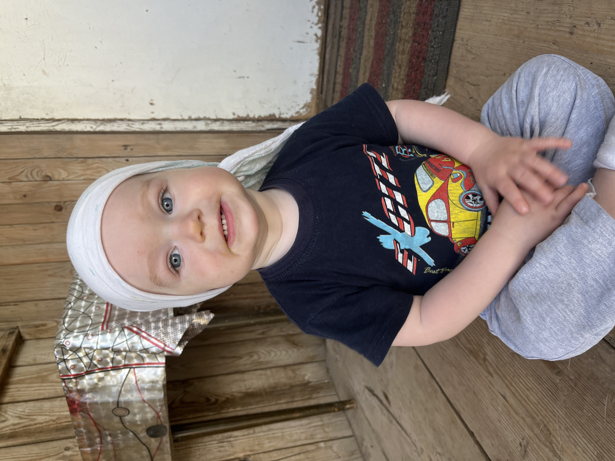 A young child with fair skin and blue eyes is seated, wearing a white headscarf, a navy t-shirt with a car design, and grey pants. The child is smiling slightly. The background includes wooden flooring and a wall, along with what appears to be a foil-covered object. The child is indoors. The location is Сосново, Russia.