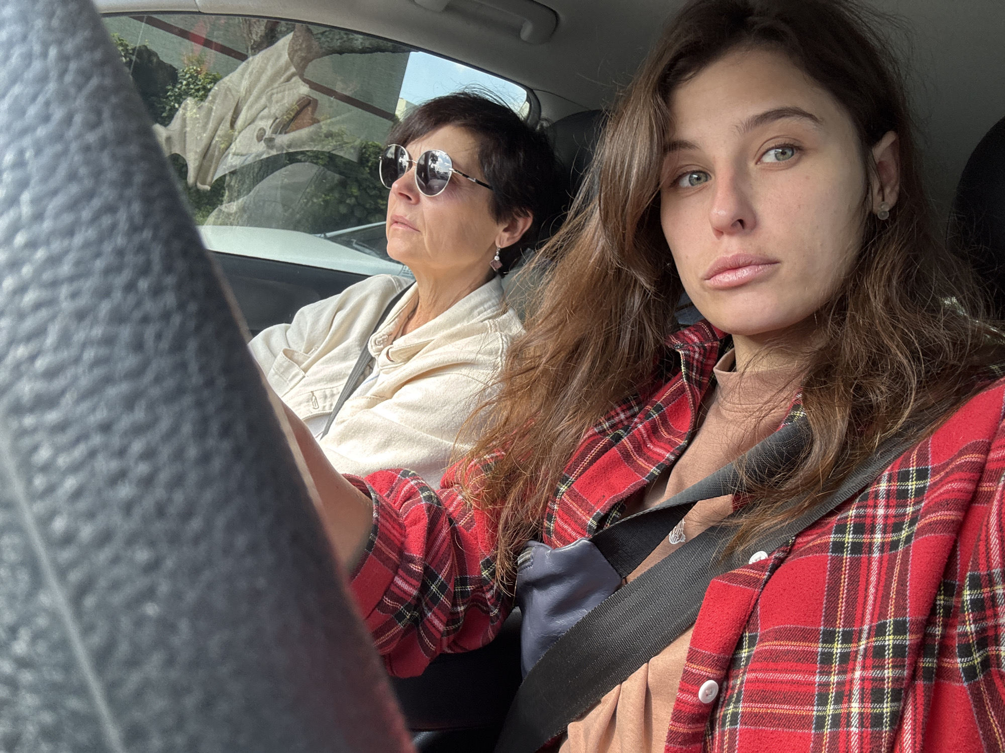 The image shows two women in a car. The woman in the driver's seat, on the left, has short dark hair and is wearing sunglasses and a light-colored shirt. The woman in the passenger seat, on the right, has long brown hair and is wearing a red and black plaid shirt over a brown shirt. Both women are wearing seatbelts. The car appears to be parked or moving slowly, and the exterior view through the windows shows greenery and buildings, suggesting an urban or suburban setting. There is no specific indication of the city of Jeju, South Korea in the image.