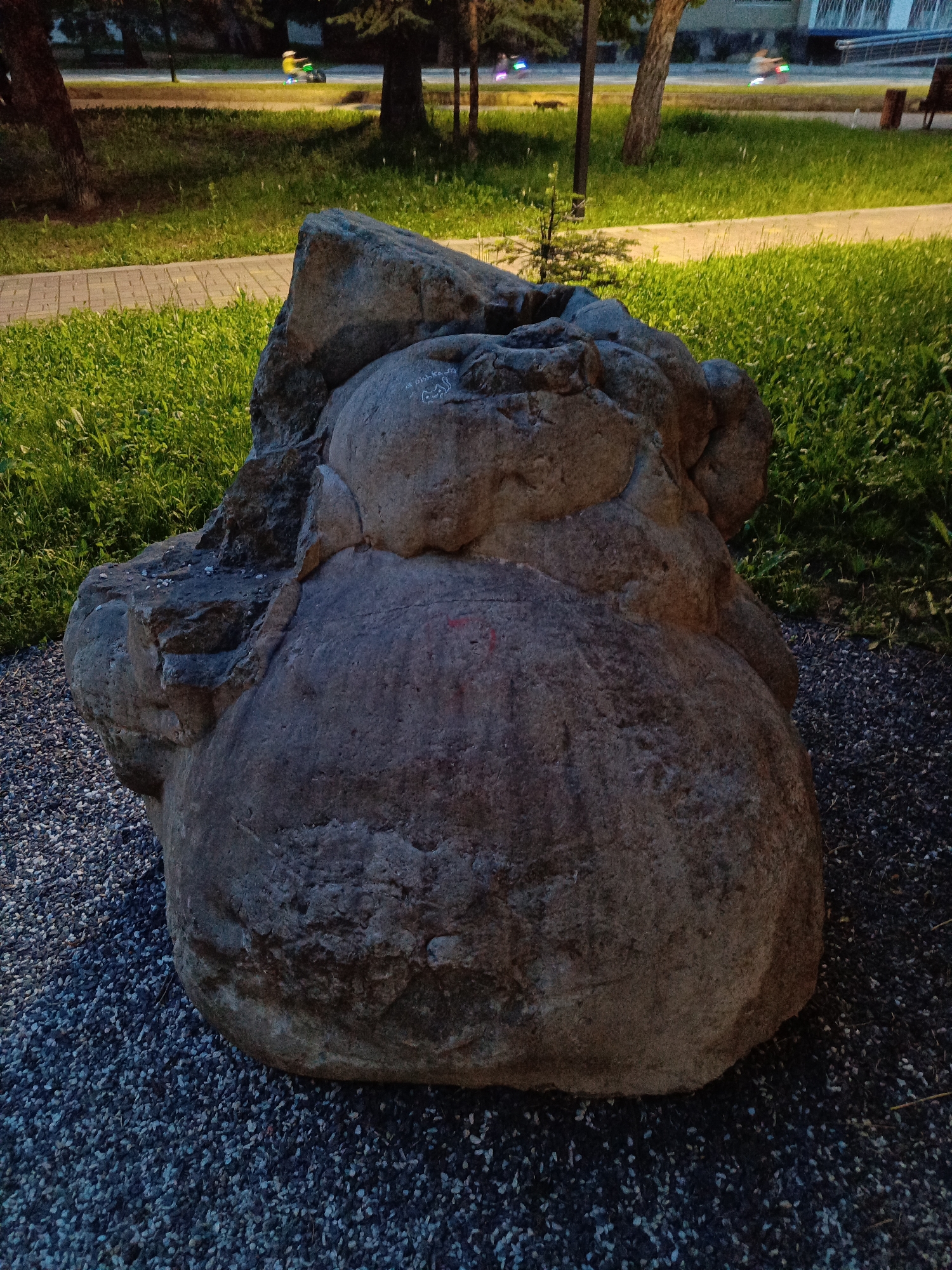 An outdoor, medium shot captures a large, weathered boulder in a park-like setting. The boulder, which appears to be a focal point, is situated on a bed of dark gray gravel. To the left of the boulder, a pathway of light-colored paving stones is visible, bordered by lush green grass. In the background, more grass and trees are present, with a paved area beyond that where two individuals are riding what appear to be electric scooters. The sky is dim, suggesting it is either early morning or late evening. A building with windows and architectural details is visible in the far background. The scene is lit by artificial lights, casting a warm glow on the grass and path. The boulder itself has some lighter markings, including what appears to be a drawing of a cat and some indistinct writing. The overall environment suggests a public urban space designed for recreation and relaxation. The location is identified as Ust-Dzheguta, Russia.