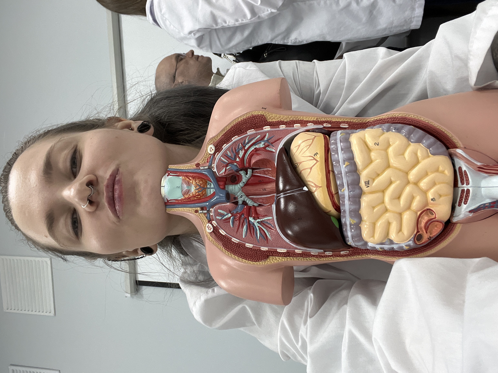 A light-skinned woman with dark hair and multiple piercings is in the foreground. She is positioned beside a mannequin of a human torso displaying internal organs. A man in a white coat is visible in the background. The environment appears to be a medical or educational setting. There is no information to indicate the location is Moscow, Russia.