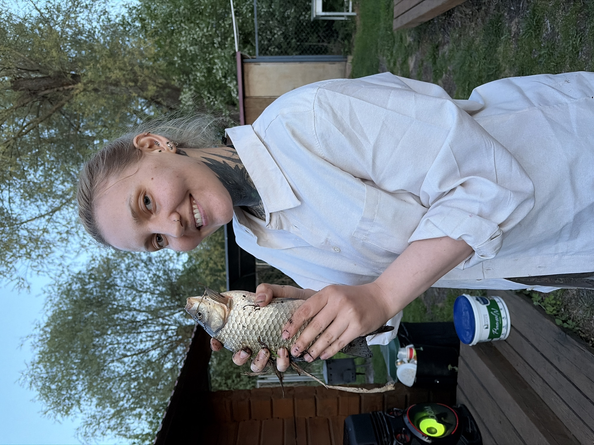 A woman with blonde hair pulled back, earrings, and neck tattoos is smiling and holding a freshly caught fish in her hands. She is wearing a white button-down shirt. The scene is outdoors, possibly in a backyard or garden setting near Malyi Bukor, Russia. In the background, there are trees, a building, and glimpses of green grass. In the foreground, there are objects on a wooden surface, including a container with a blue lid and some dark equipment.