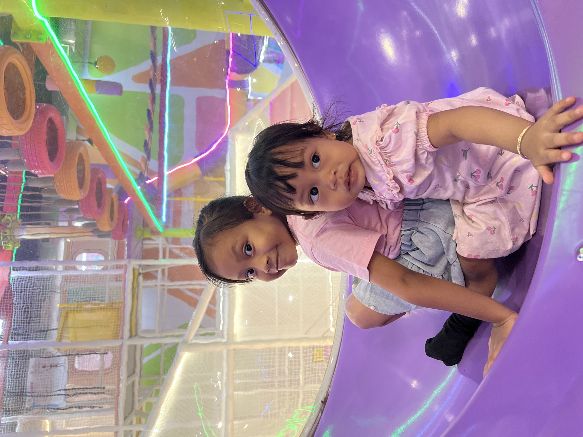 Two young girls are sitting on a purple slide in an indoor playground in Kecamatan Pati, Indonesia. The girl on the left is older, wearing a pink t-shirt and denim shorts. She is smiling and looking to her left. The girl on the right is younger, wearing a pink dress with a cherry pattern. She is looking forward with a slightly surprised expression. The background is a colorful and complex structure of the playground, with netting, tubes, and bright lights. The lighting is mixed, with some areas well-lit and others in shadow, suggesting an indoor artificial lighting environment.
