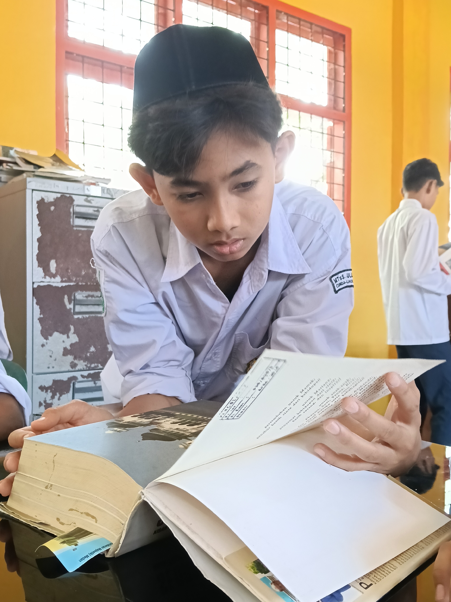 This indoor photograph features a young male student deeply engrossed in reading a large book. He is positioned centrally, wearing a white long-sleeved uniform shirt and a black songkok (a traditional cap worn by Muslim men). His gaze is directed intently at the open pages of the book, which rests on a dark surface, likely a table. His right hand carefully holds open another section of the book.
On the student's right sleeve, a visible patch reads "MTSS ULL CUNDA-LHOC," indicating he is a student of Madrasah Tsanawiyah Swasta Ulul Albab Cunda, an Islamic junior high school in Lhokseumawe, Indonesia.
In the background, another male student in a white shirt is partially visible on the right, also appearing to be reading. To the left, an old, weathered metal filing cabinet with peeling paint suggests an established, possibly aged, educational setting. The room has bright yellow walls and windows with prominent red frames and grilles, through which bright daylight streams, indicating it is daytime.
Visible text on the book pages includes fragments of Indonesian phrases such as "Persembahan untuk Mamak" (Dedication for Mother), "kehidupan," and "kasih sayang," along with "Majestik) Medan" and "PENGOL." The overall scene depicts a focused learning environment at an Islamic school in Lhokseumawe, Indonesia.