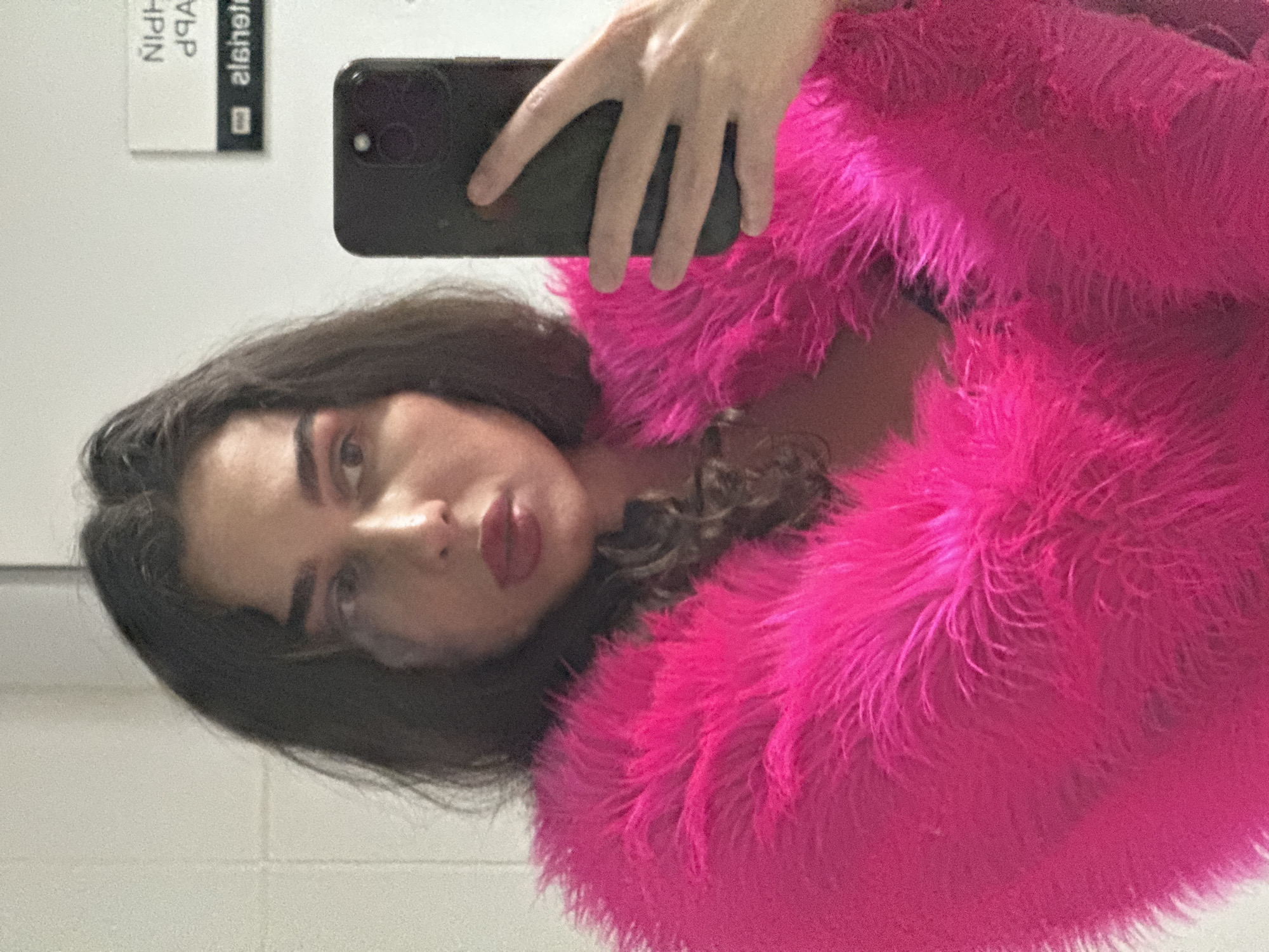 A woman with dark hair and makeup takes a selfie in a brightly lit room with tiled walls. She is wearing a fluffy hot pink jacket over a dark garment. She is holding a black smartphone in her right hand, angled to capture her reflection in a mirror. In the background, a sign with mirrored text is visible on the wall. The location is a public restroom in Kazan, Russia.