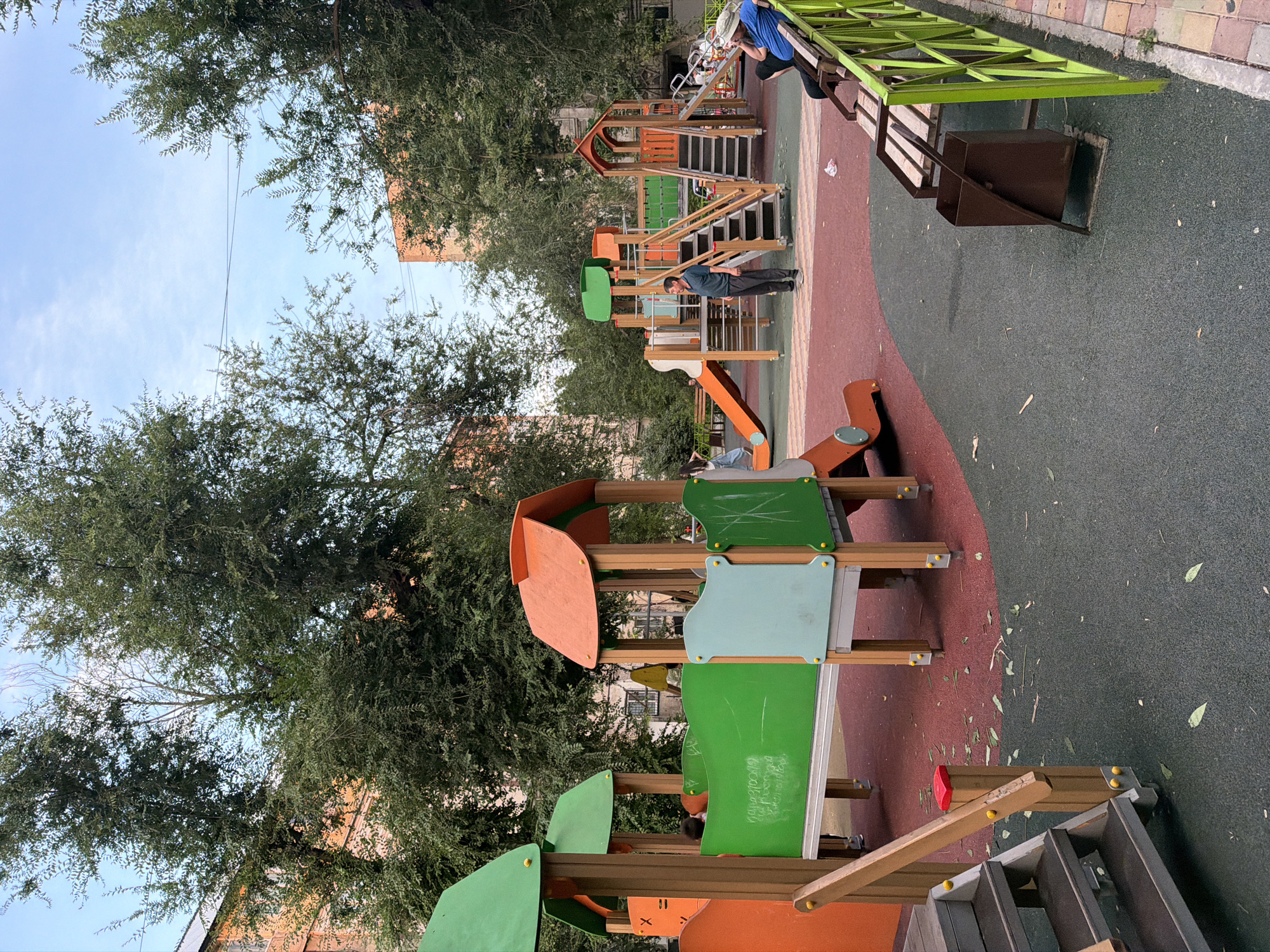 The image depicts a vibrant, modern outdoor playground situated in an urban courtyard setting in Shymkent, Kazakhstan. Tall, mature trees with dense green foliage flank the playground, casting dappled shade on the scene. Buildings with orange brick facades are visible through the trees, suggesting a residential area. The playground itself features colorful structures, including slides, climbing frames, and platforms, all constructed from wood and plastic. The ground surface is a combination of red and dark green rubberized material, designed for safety.
In the foreground, a man in a dark blue polo shirt and gray pants stands near the center of the playground, looking towards the structures. To the right, a wooden bench is occupied by two individuals. One person, wearing a blue shirt and a hat, is seated, while another, in a blue jacket and hat, leans back, appearing relaxed. A brown trash receptacle stands near the bench.
Several children are present in the playground. A person with dark hair, possibly a child, is partially visible behind a green barrier on one of the play structures. Another person, appearing to be a young woman with light-colored clothing, is walking away from the playground on the left side of the image. Further back, a child is seated on a slide.
The overall impression is one of a well-maintained, active community space within a city environment. The sky above is partly cloudy, indicating daytime. The presence of fallen leaves on the ground suggests it might be autumn.