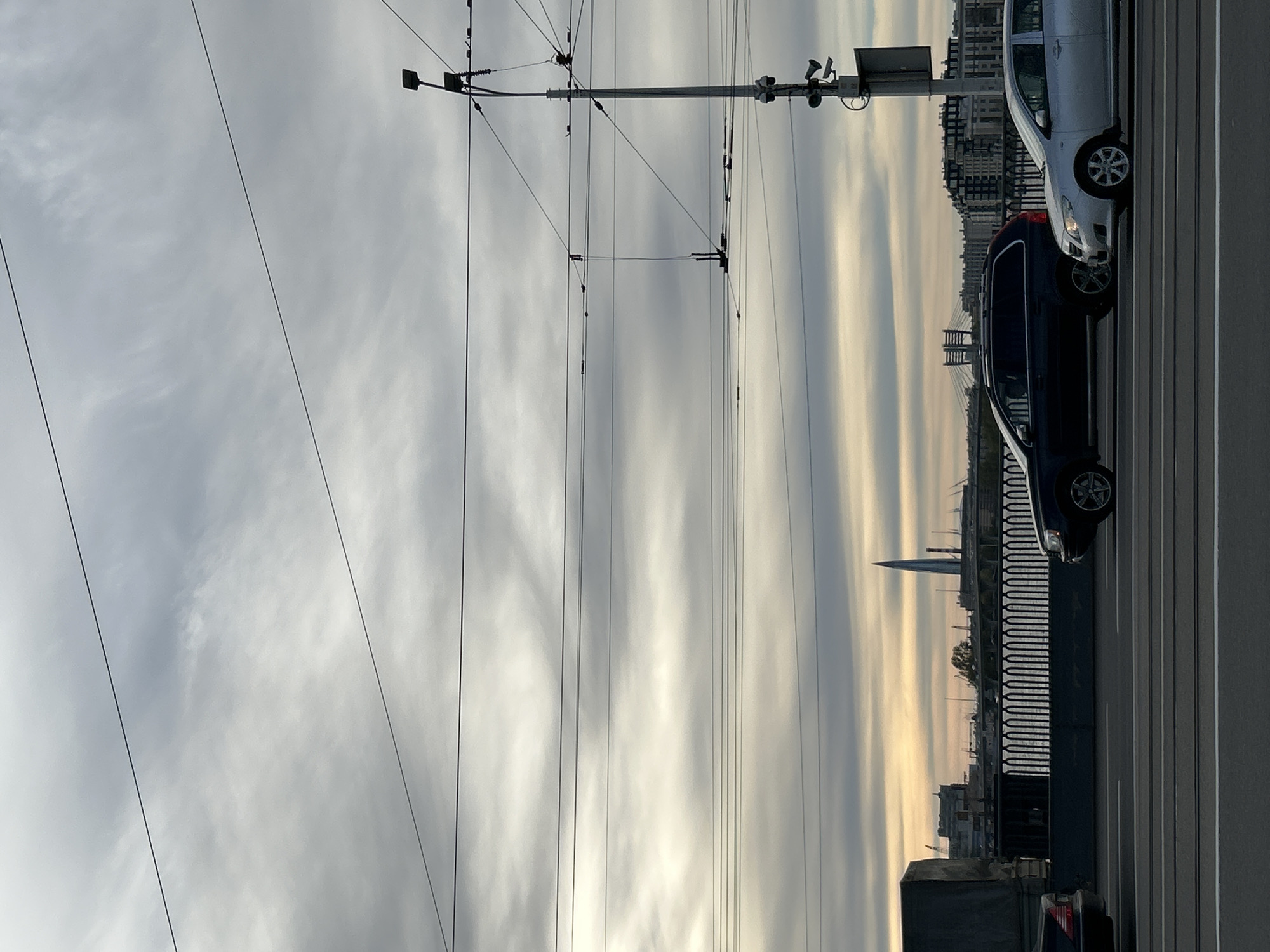 The image shows a view from a bridge in Saint Petersburg, Russia. The sky is overcast with layered clouds. Tram wires run across the sky. A dark-colored car and a silver car are parked on the right side of the frame. A bridge can be seen in the distance, along with the Lakhta Center skyscraper. A metal fence runs along the side of the bridge.