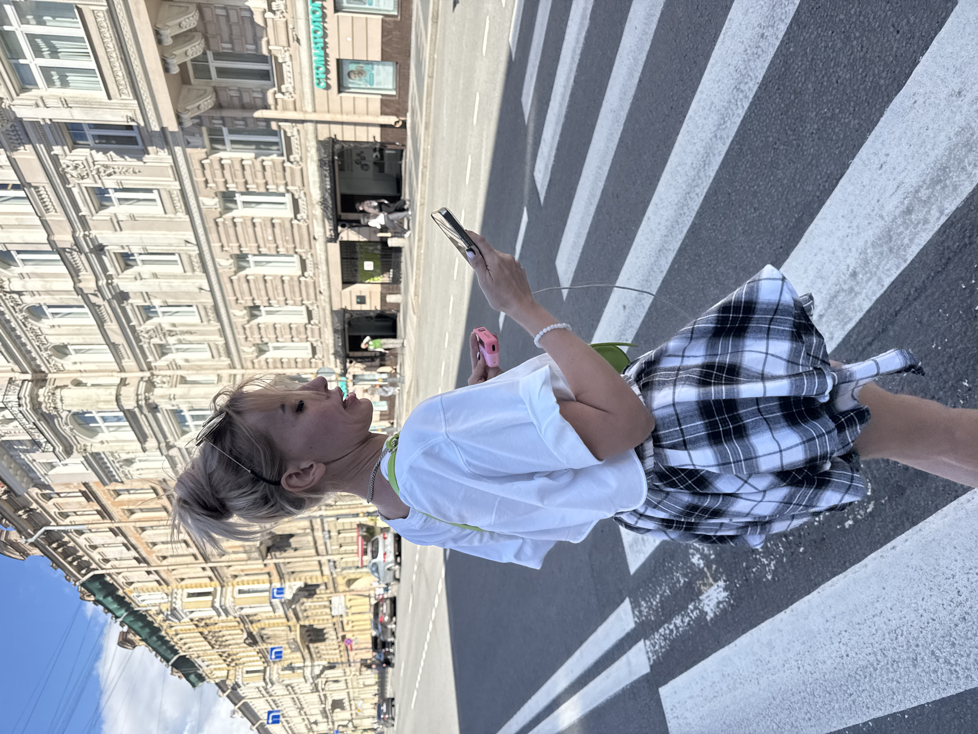 The media file shows a young woman walking across a pedestrian crossing in Saint Petersburg, Russia. She has blonde hair pulled back into a bun and is wearing sunglasses on top of her head. Her outfit consists of a white t-shirt and a black and white plaid skirt. She is holding a smartphone in her right hand and a pink object, possibly a portable charger or case, in her left hand. A green strap, likely from a bag, is visible around her shoulder.
The scene is set on a city street with an ornate, multi-story building on the left side of the image, characteristic of Saint Petersburg's architecture. The building has many windows and decorative elements. The pedestrian crossing is clearly marked with white stripes on an asphalt road. Several cars are visible in the background, suggesting it is daytime and the street is in use. The sky appears blue with some clouds, indicating fair weather. The overall mood seems casual and everyday, with the woman appearing engaged with her phone as she crosses the street.
There is visible text on the building, including "СТОМАТОЛОГ" which translates to "Dentist". Other signage is present but not clearly legible.