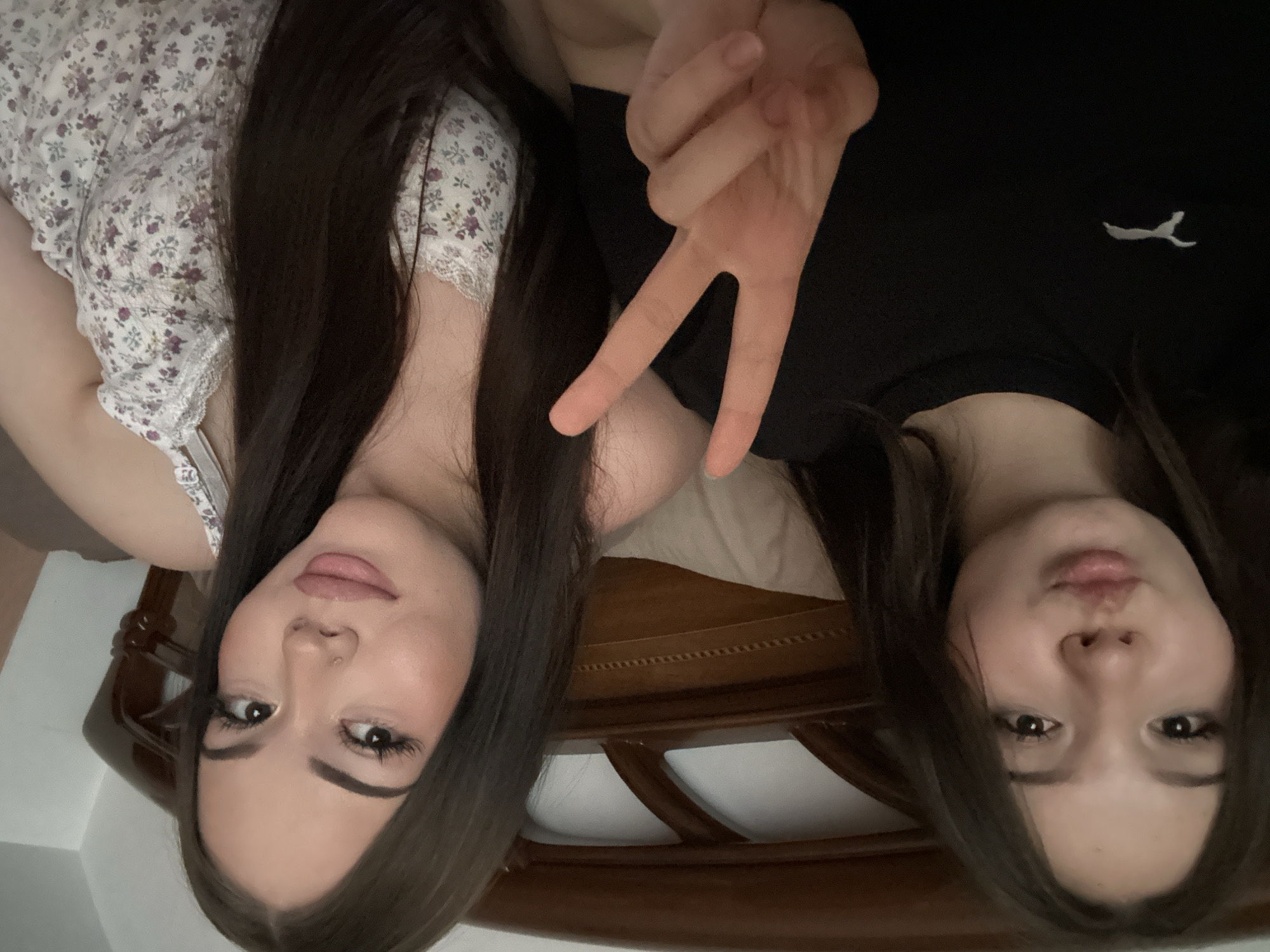 The image, taken in Naberezhnye Chelny, Russia, shows two young women lying down, looking upwards at the camera. The perspective suggests they are on a bed, with a dark wooden bed frame partially visible in the background between them.
The woman on the left has long, dark hair and is wearing a white top with a delicate purple floral pattern and white lace trim. She has noticeable makeup, including dark eyebrows, prominent eyeliner, and light pink lipstick. A small nose piercing is visible on her left nostril.
The woman on the right also has long, dark hair and is wearing a plain black top. A small white embroidered logo, identified as the Puma leaping cat, is visible on the left side of her chest. Her hand is raised in the center of the frame, making a peace sign gesture. She also wears makeup, with dark eyebrows, eyeliner, and light pink lipstick, and has a slightly pursed-lip expression.
Both individuals appear relaxed and are posing for the photograph, which seems to have been taken indoors with soft lighting.
