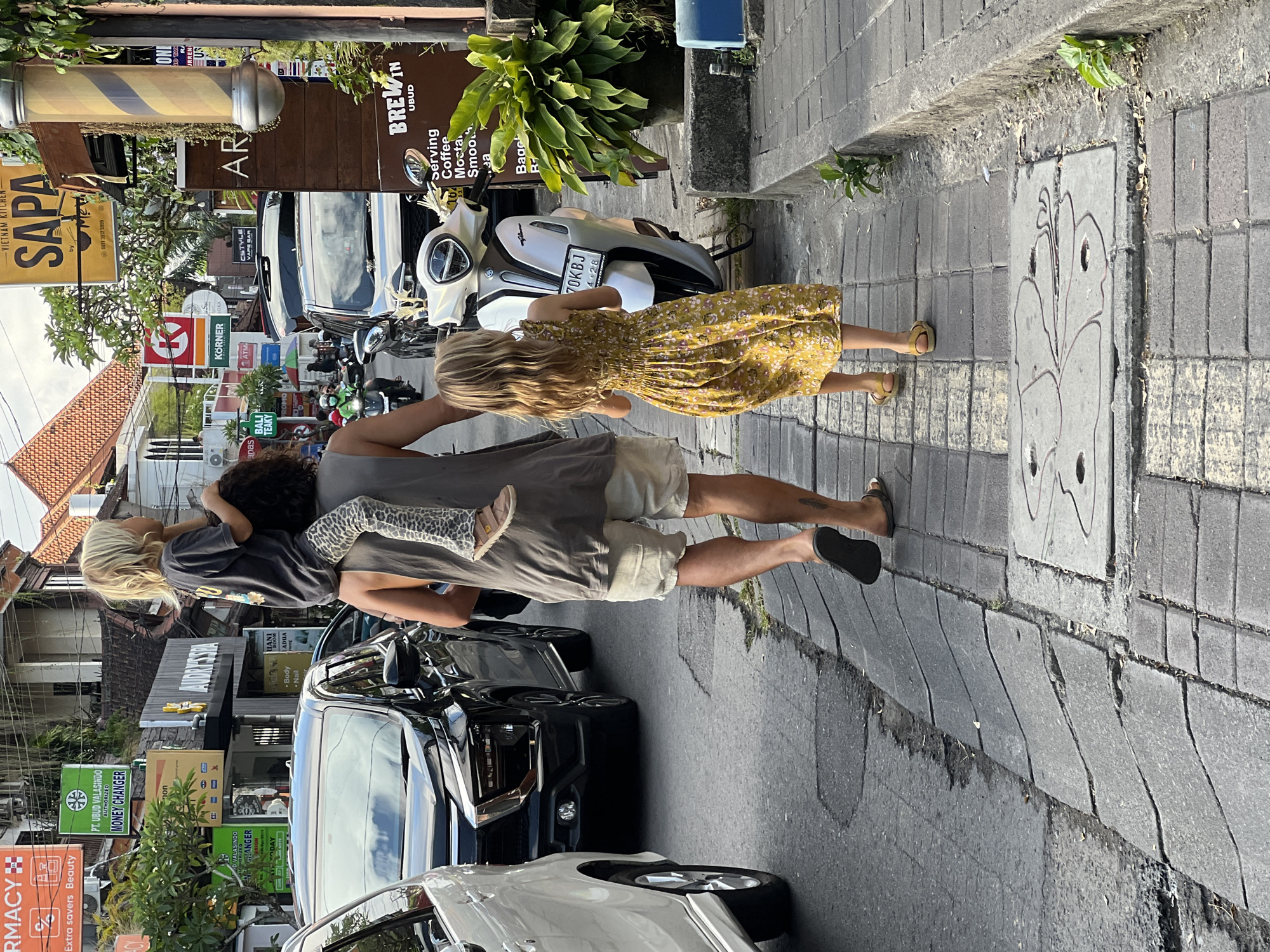 The image depicts a street scene in Ubud District, Indonesia. A man carries a child on his shoulders while another child walks alongside him. The street is lined with shops and scooters, a parked car, and various commercial signs, including "SAPA - VIETNAM KITCHEN" and "PT. UBUD VALASINDO MONEY CHANGER." A drain cover with a flower drawing is visible on the pavement. A barber's pole sign can be seen in the background, along with a restaurant sign that reads "BREWIN UBUD."