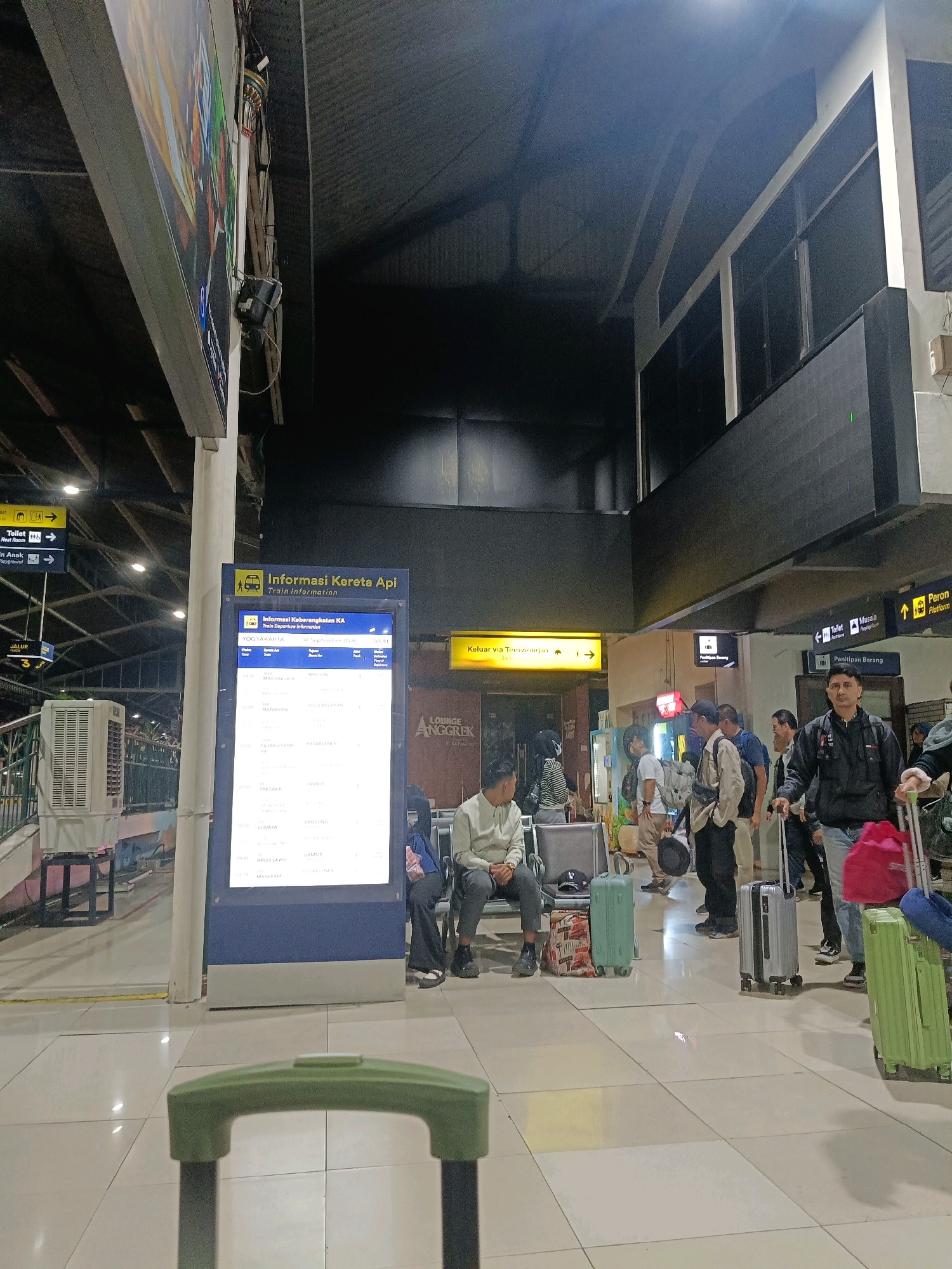 The image depicts an indoor scene at a train station, likely Yogyakarta Tugu Station in Yogyakarta, Indonesia, given the signage. The perspective is low, focusing on the lower half of a large green suitcase in the foreground. Behind the suitcase, the polished floor of the station is visible, reflecting the ambient light.
In the middle ground, several individuals are present. One man in the foreground, facing the camera, wears a black jacket and jeans and is holding a suitcase handle. To his left, a silver rolling suitcase stands upright. Further left, a woman in dark clothing is pulling a bright pink bag on wheels. Beside her, a man in a black jacket is speaking to another man in a light-colored shirt and dark pants. Several other people are standing or sitting on benches in the background.
A large, illuminated digital display board with train information stands prominently in the center-left of the frame. The board shows train departures and arrivals, with "Informasi Kereta Api" (Train Information) and "Informasi Keberangkatan KA" (Train Departure Information) as headers. To the left of this board, a white air conditioning unit is visible.
The background reveals the architecture of the station, with a dark, corrugated ceiling and exposed beams. Various directional signs are present, including one for "Toilet," "Penitipan Barang" (Luggage Storage), and a yellow sign with an arrow indicating an exit. The overall atmosphere suggests a busy transportation hub.